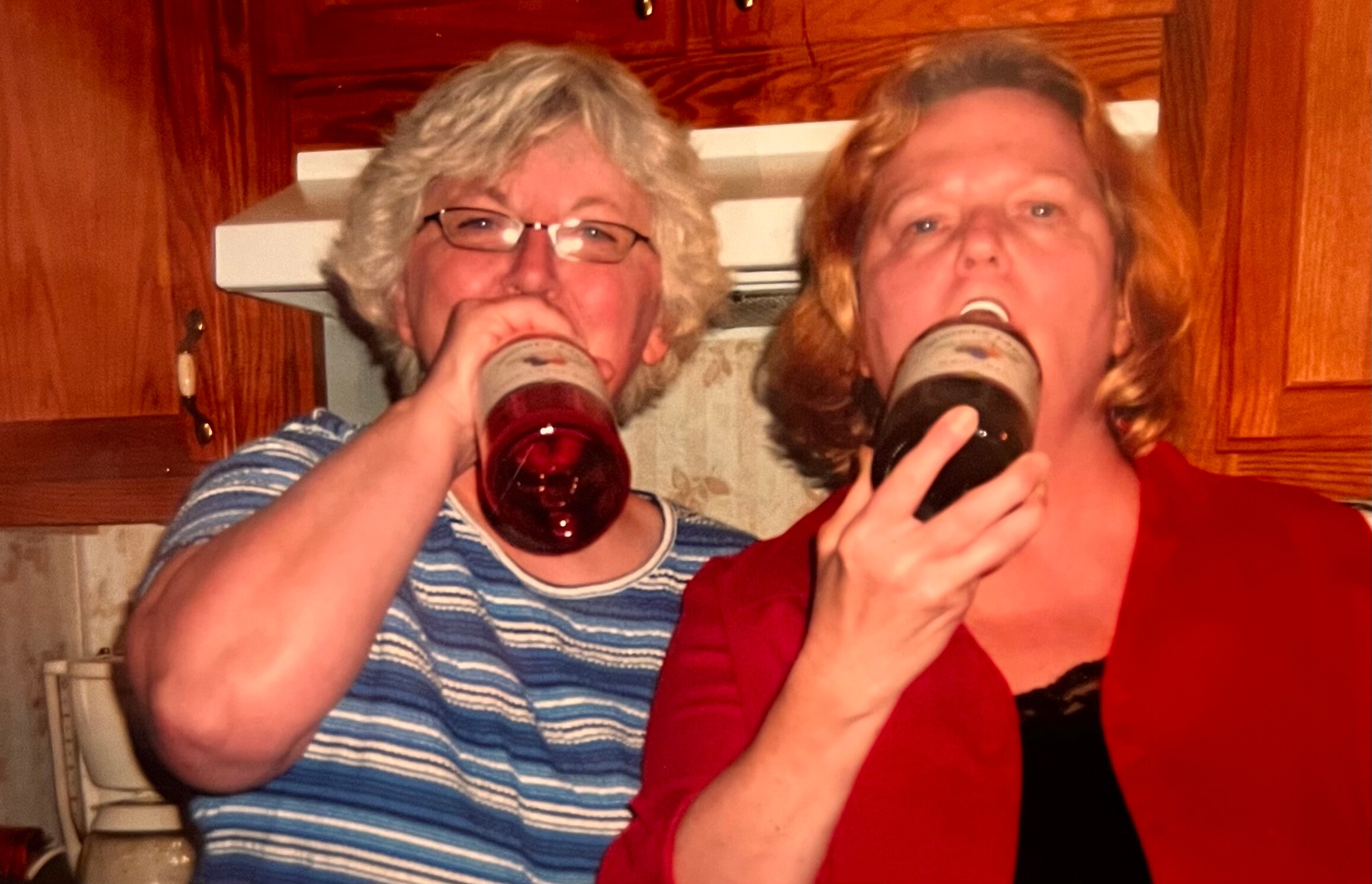 Two women drink beer from bottles in a home kitchen, smiling at the camera.
