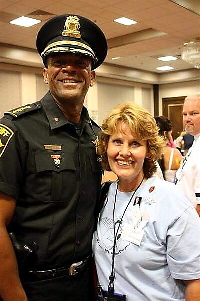 Police officer in dress uniform posing with a smiling woman in a light blue shirt at an indoor event.