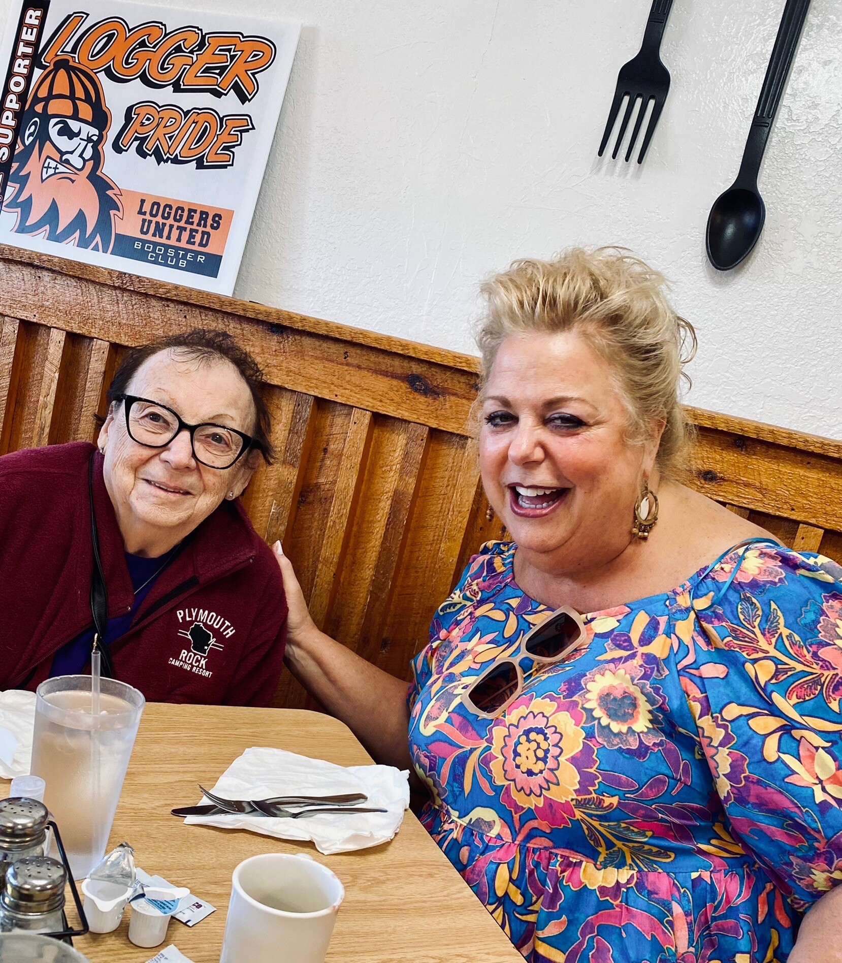 Two women smile for a selfie at a casual restaurant table with cups and utensils, wood-paneled bench behind them and a 'Logger Pride' poster on the wall behind the left person.