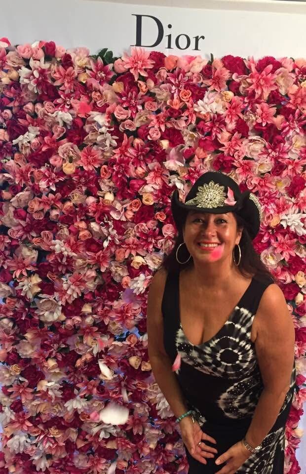 Woman smiling in front of a pink flower wall with 'Dior' sign, wearing a black hat with a jeweled centerpiece and a black-and-white dress.