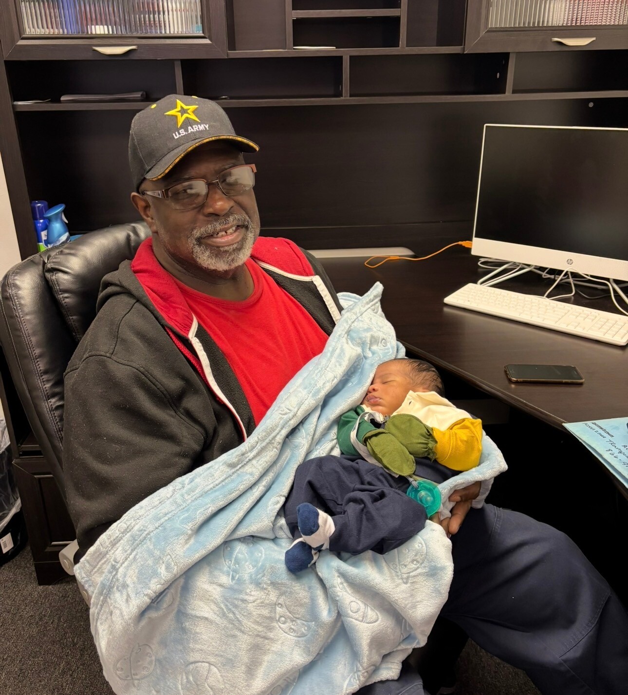Man wearing a U.S. Army cap and red shirt sits in an office chair, cradling a sleeping newborn wrapped in a light blue blanket at a desk with a computer nearby.