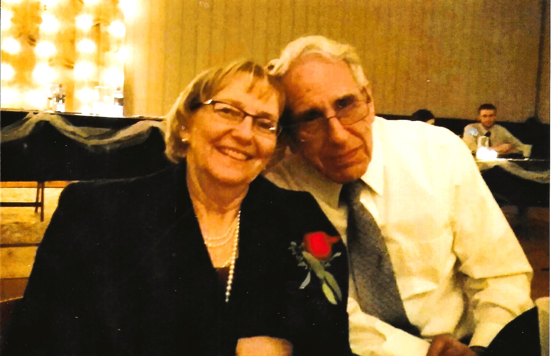 Two smiling older adults posing closely for a photo at a formal event, with a red boutonniere on the woman's lapel and warm lighting behind them.