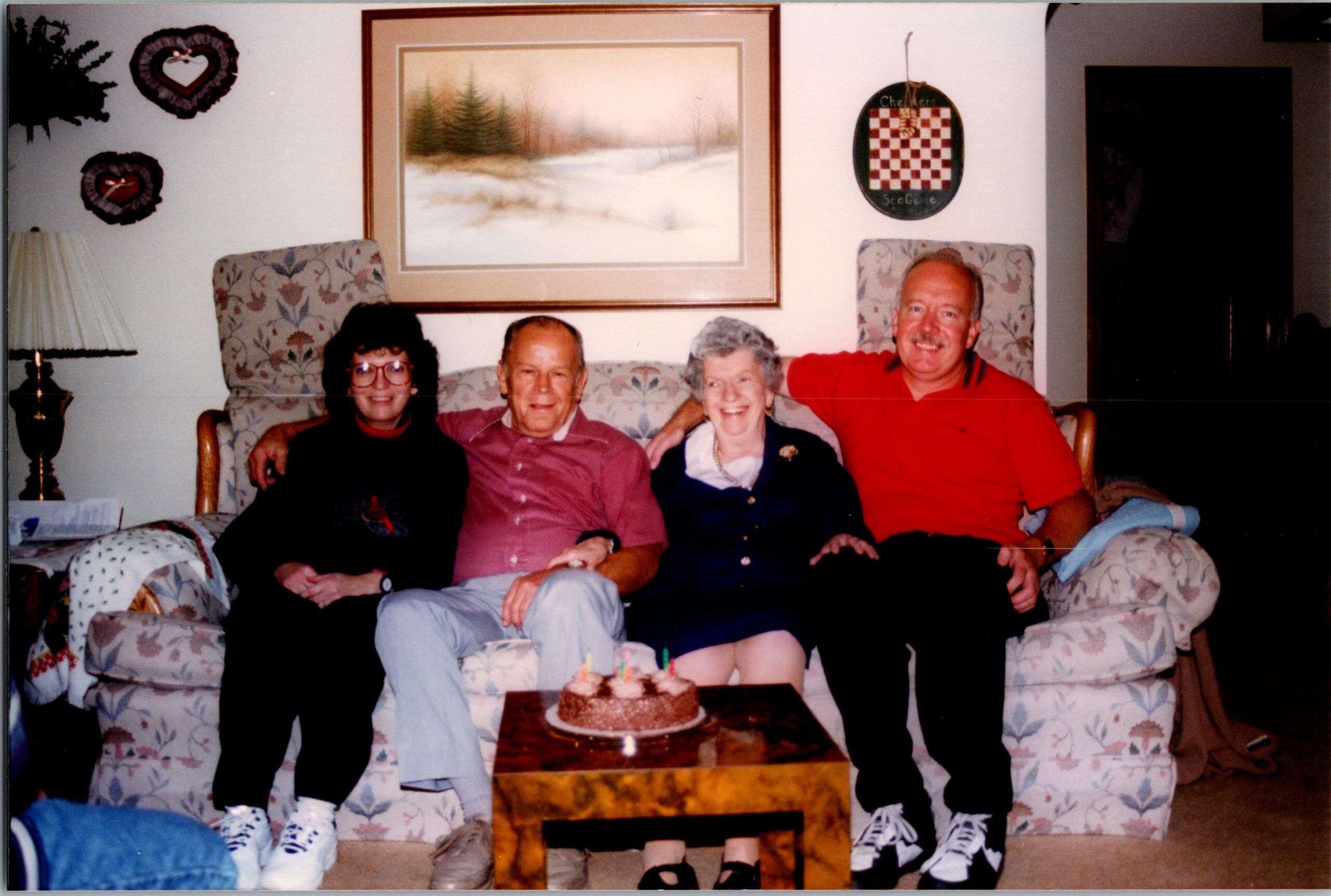 Four adults sit on a floral couch in a living room, smiling, with a small chocolate cake on the coffee table.