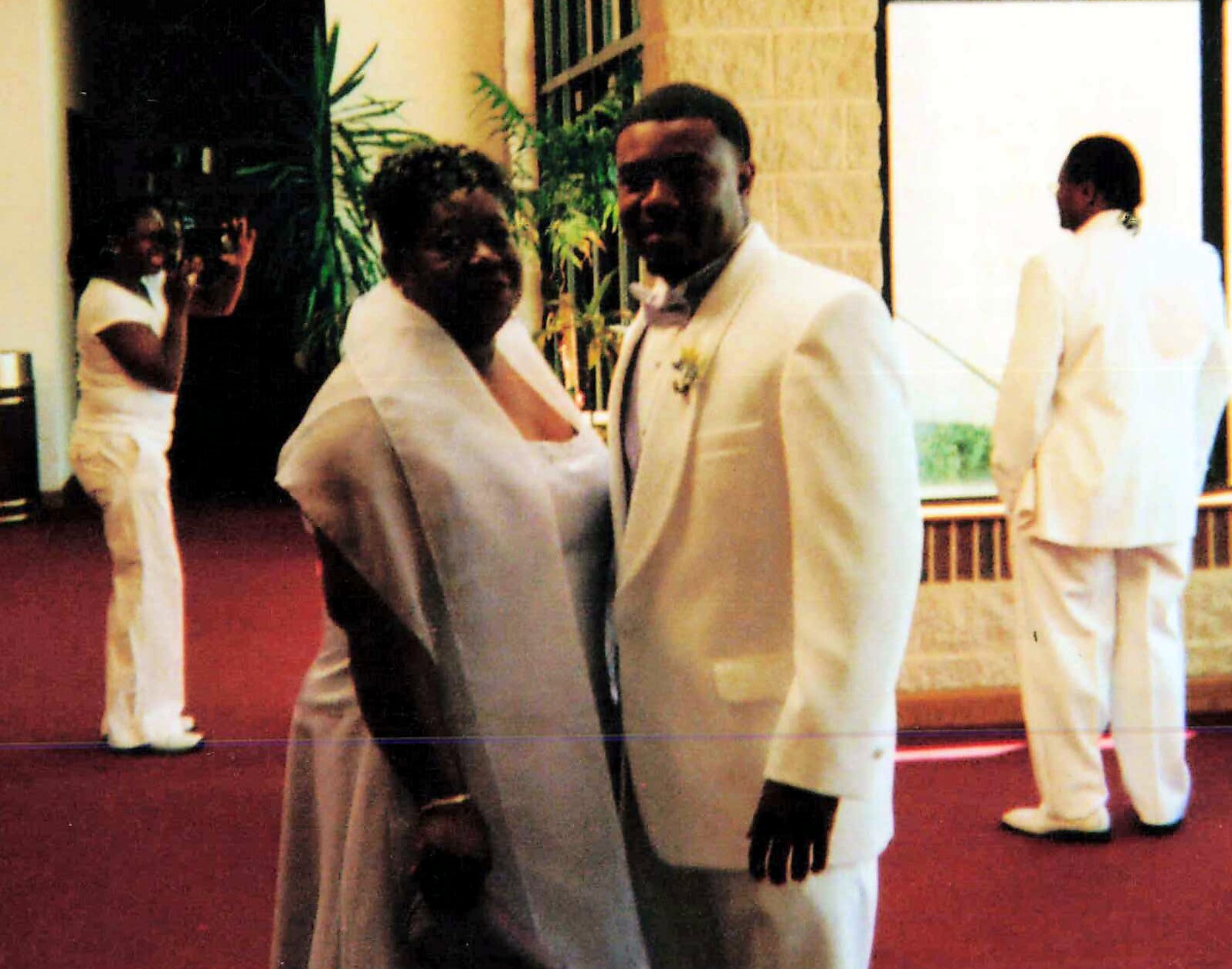 Couple in formal white attire posing indoors on a red carpet with greenery in the background.