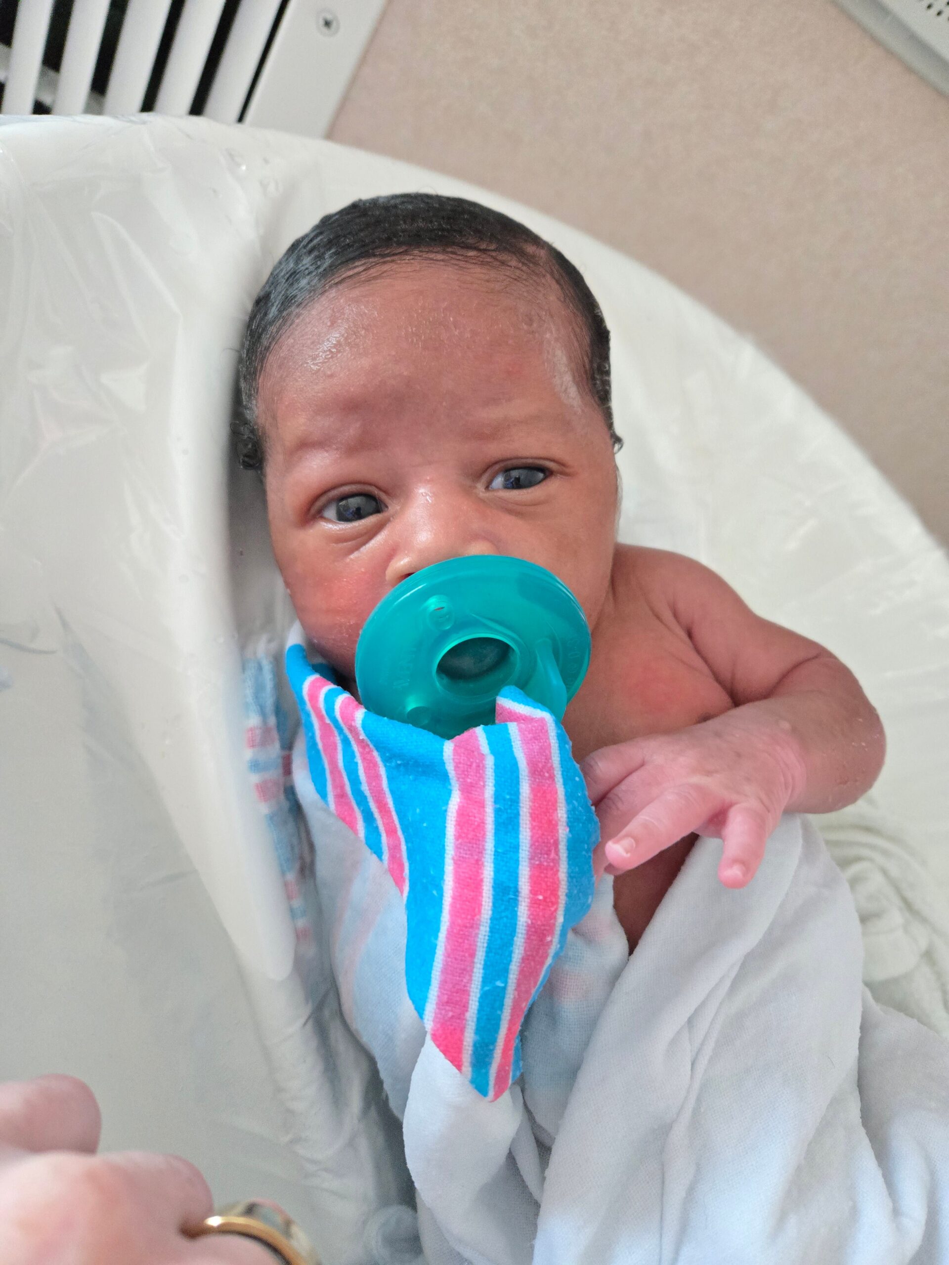 Newborn baby with blue-green pacifier, wrapped in a striped blue and pink bib, resting in a light blanket in a hospital setting.