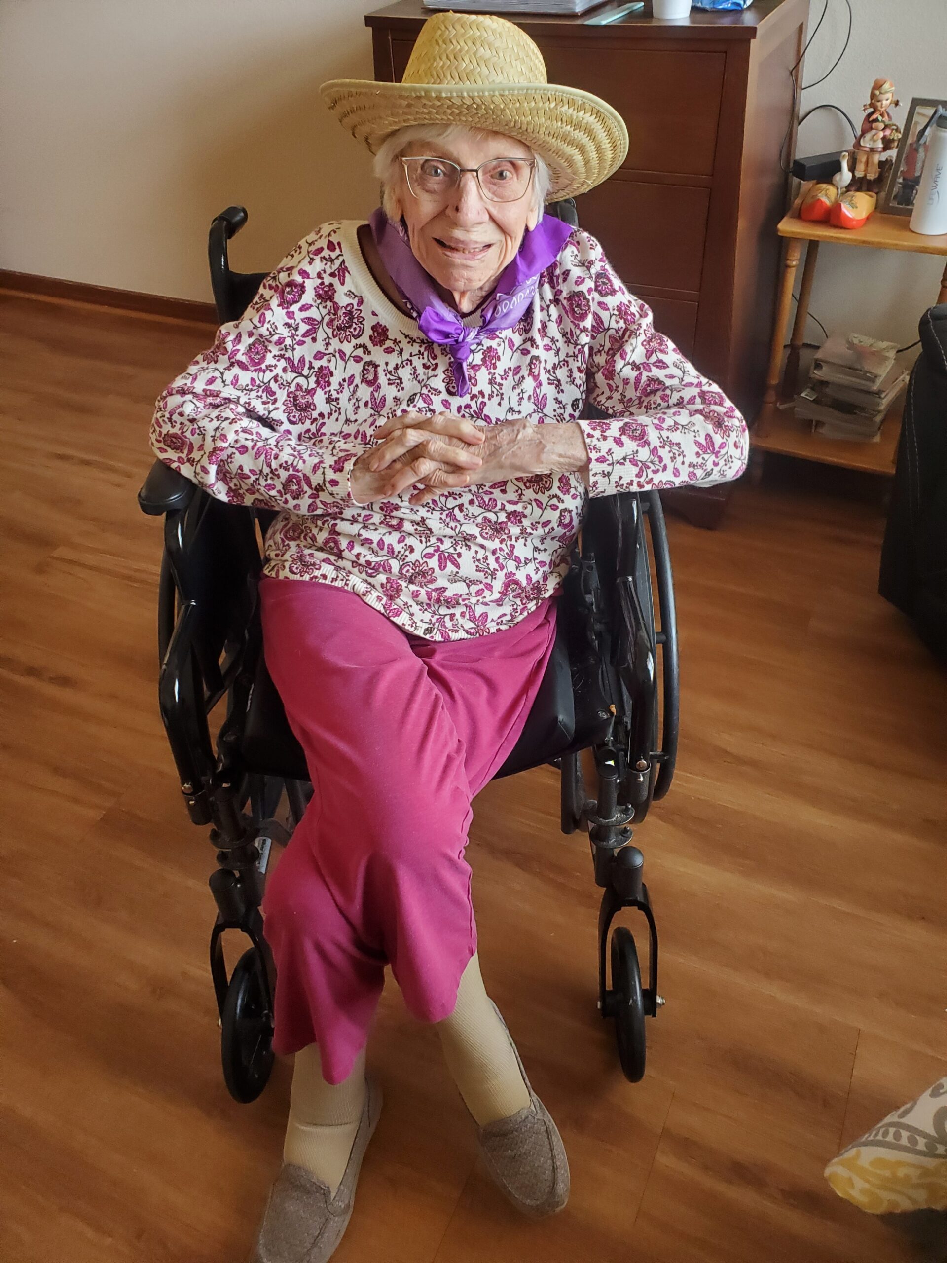 Elderly woman in a wheelchair wearing a straw hat, floral sweater, and purple scarf, smiling at the camera in a living room.