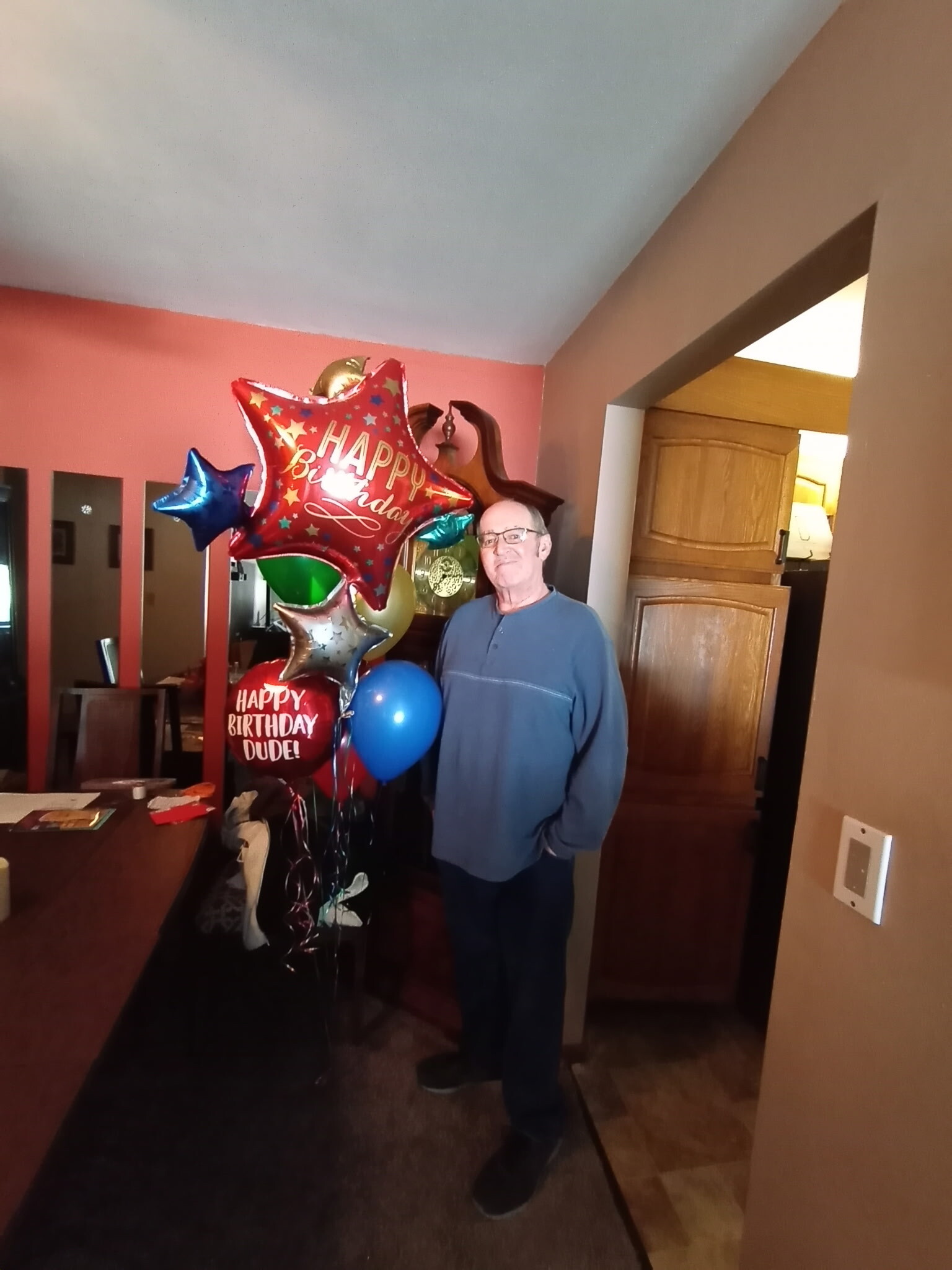 Older man in a blue sweater stands in a home hallway, surrounded by colorful birthday balloons with a red star-shaped balloon saying 'Happy Birthday'