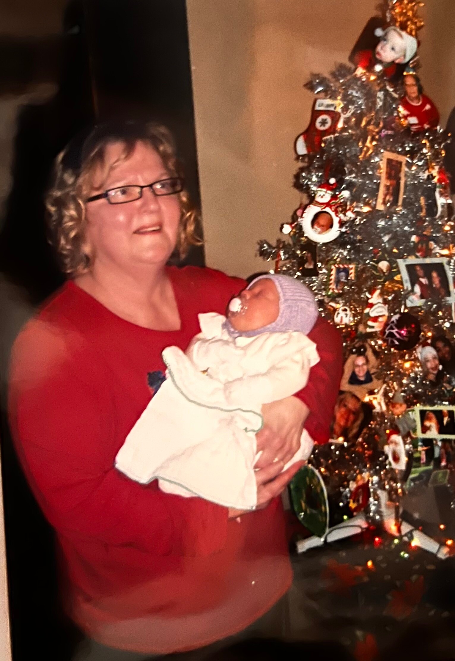 Woman in a red sweater holding a newborn in front of a decorated Christmas tree.