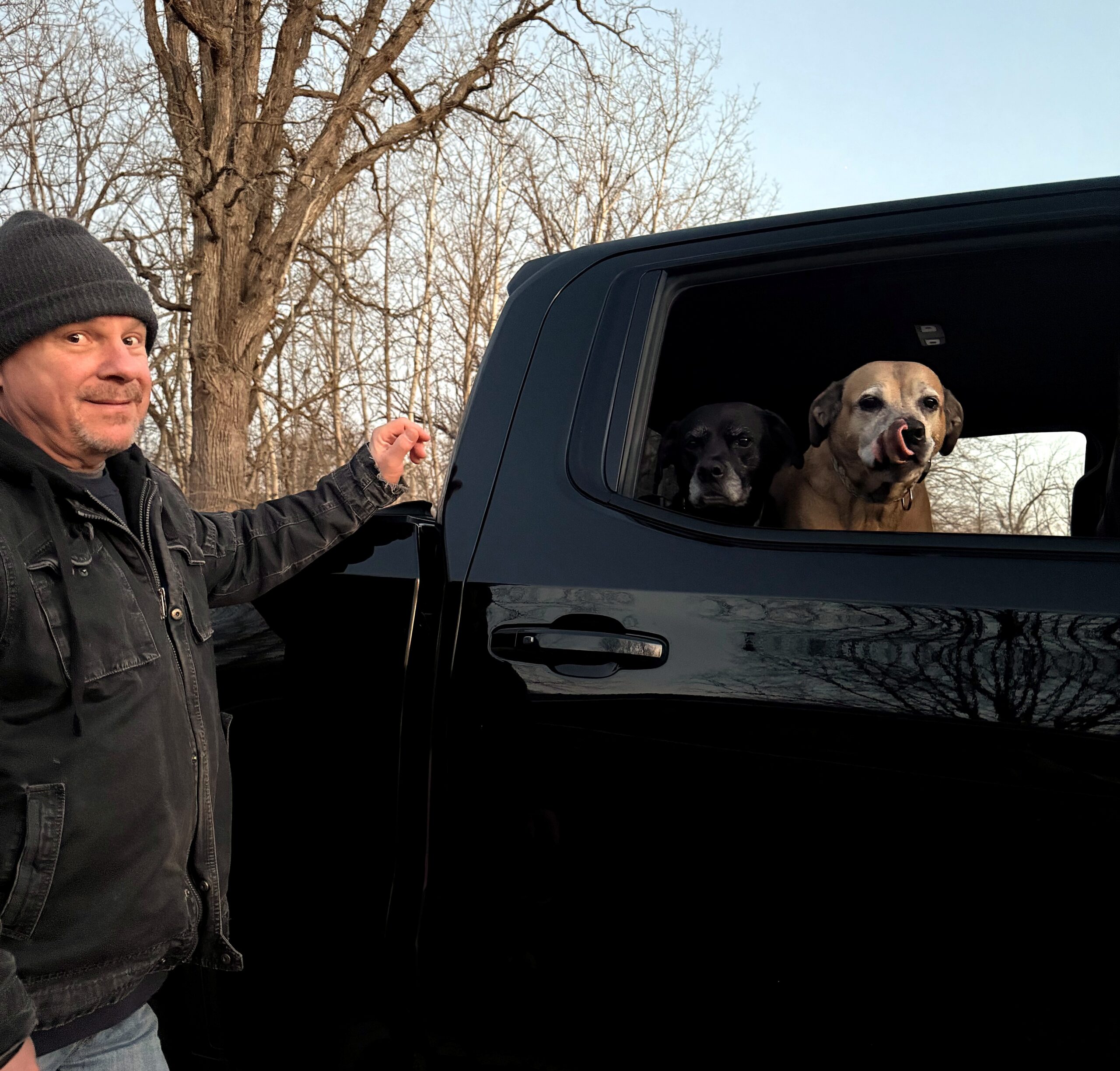 Man in a dark jacket and gray beanie stands beside a black SUV, with dogs visible in the rear seat window awaiting a ride.