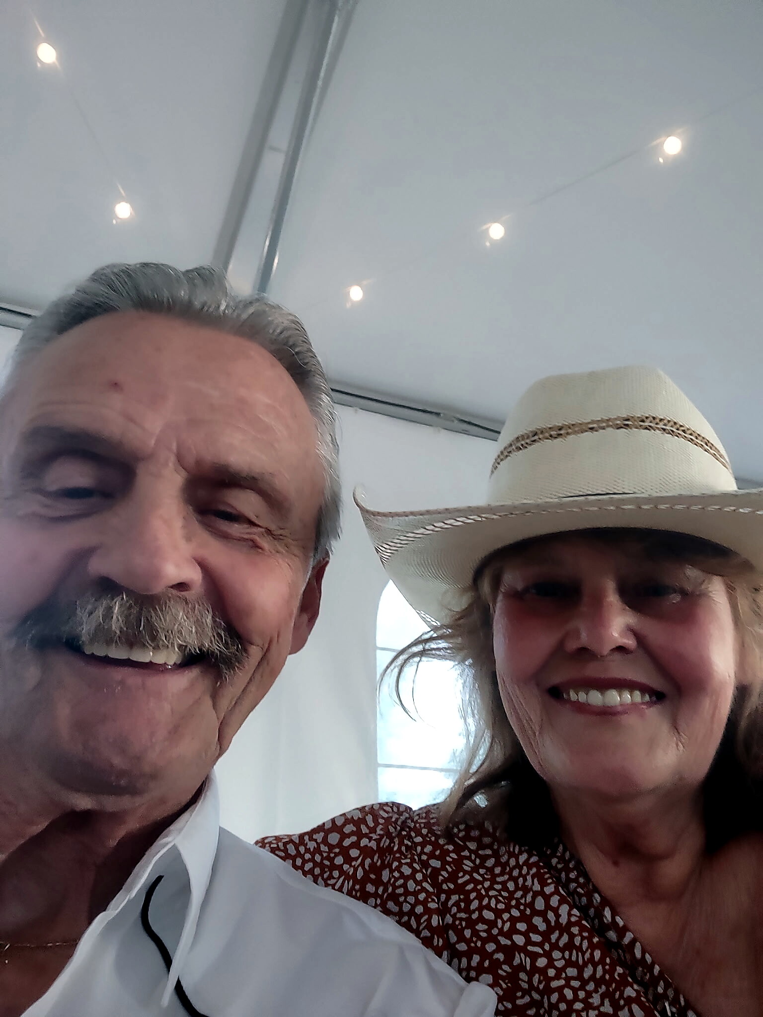Two people smiling for a selfie inside a tent or canopy, with string lights overhead; man on the left has gray hair and a mustache, woman on the right wears a wide-brim hat.