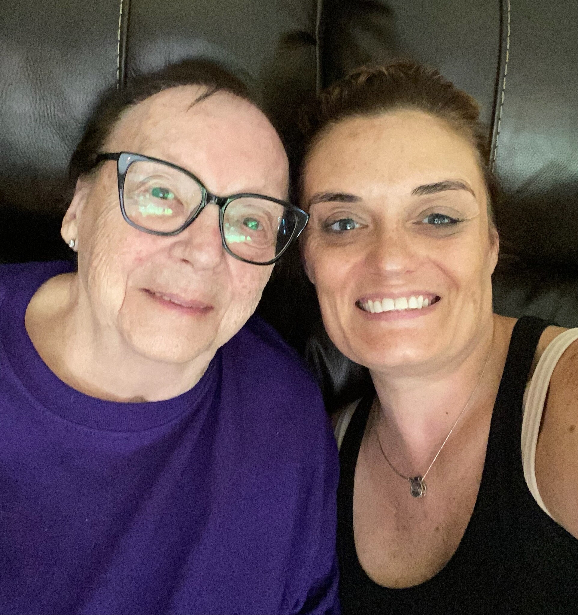 Two women taking a close-up selfie, smiling at the camera; one wearing glasses and a purple shirt, the other in a black top with a necklace inside a car-like background.