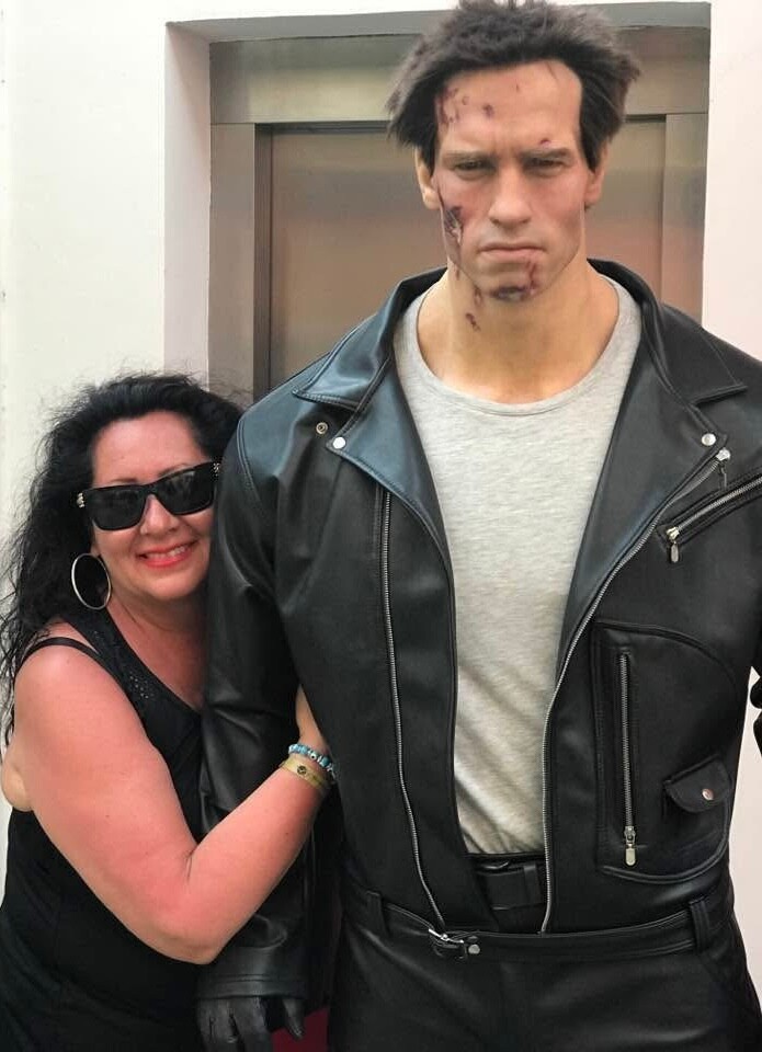 Smiling woman in black clothes and sunglasses hugs a tall man in a black leather jacket with torn-face makeup.
