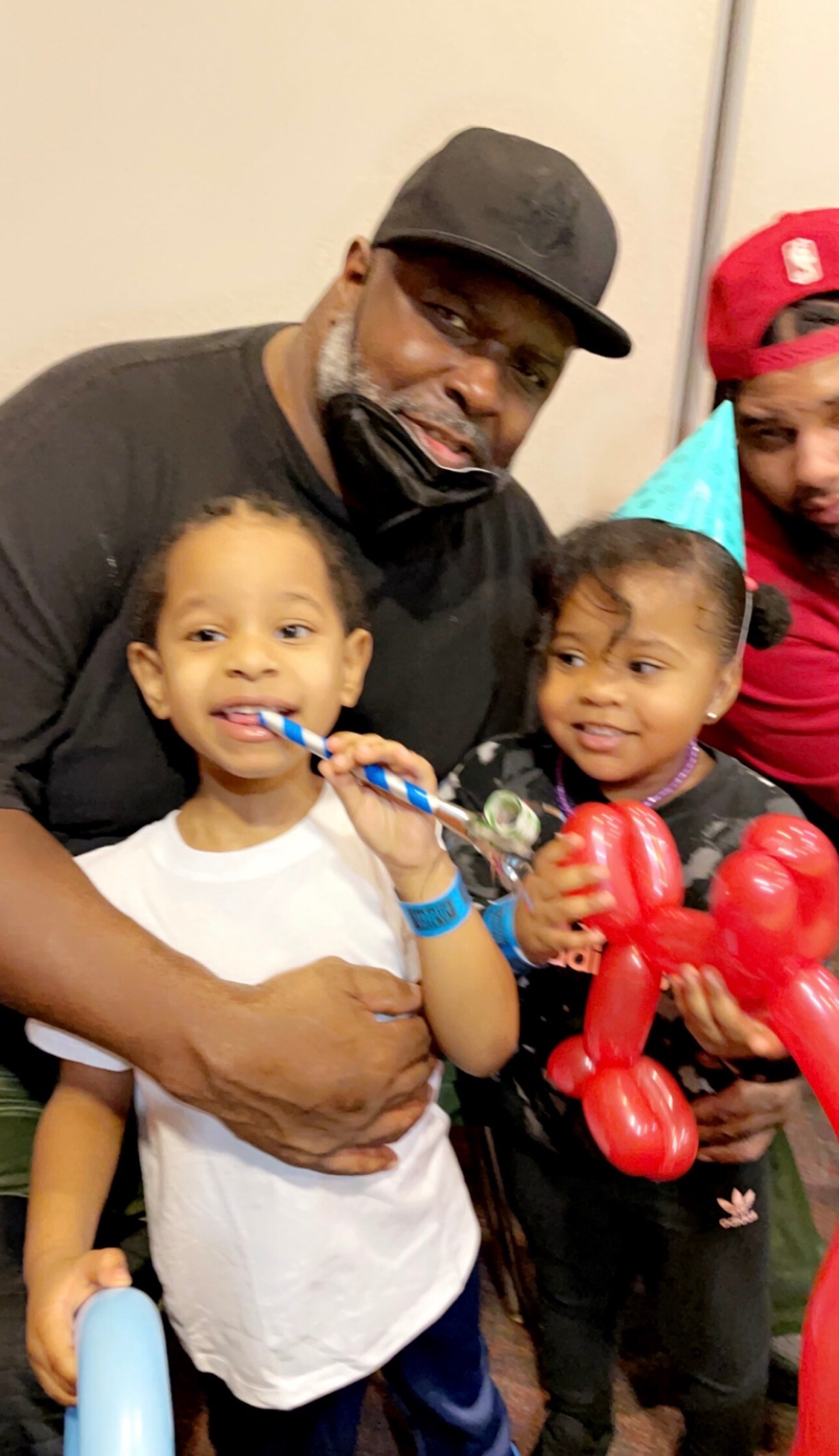 A man in a black cap and shirt holds two smiling kids at a party, one with a blue party horn and the other with red balloon animals in her hands.