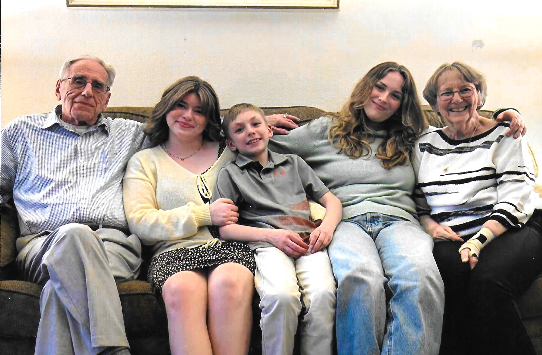 Five family members sitting close together on a couch, smiling at the camera: an elderly man, a young woman, a boy, another young woman, and an elderly woman on the right.