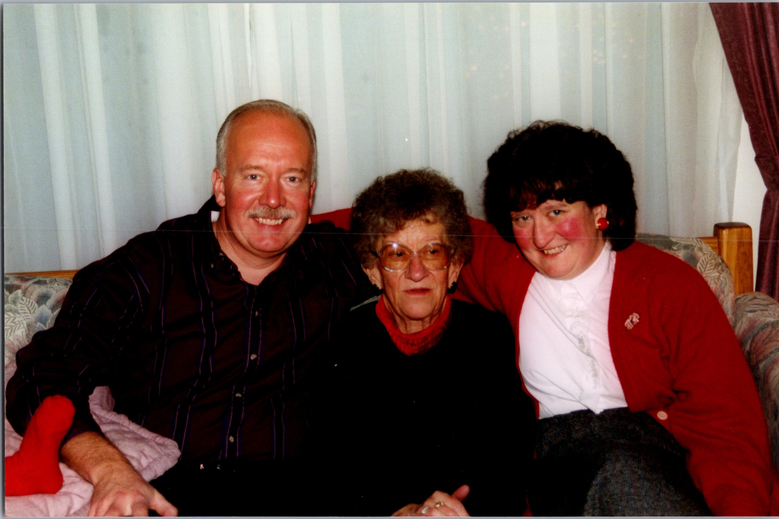 Three adults sitting close together on a sofa, smiling at the camera in a living room setting.] ,