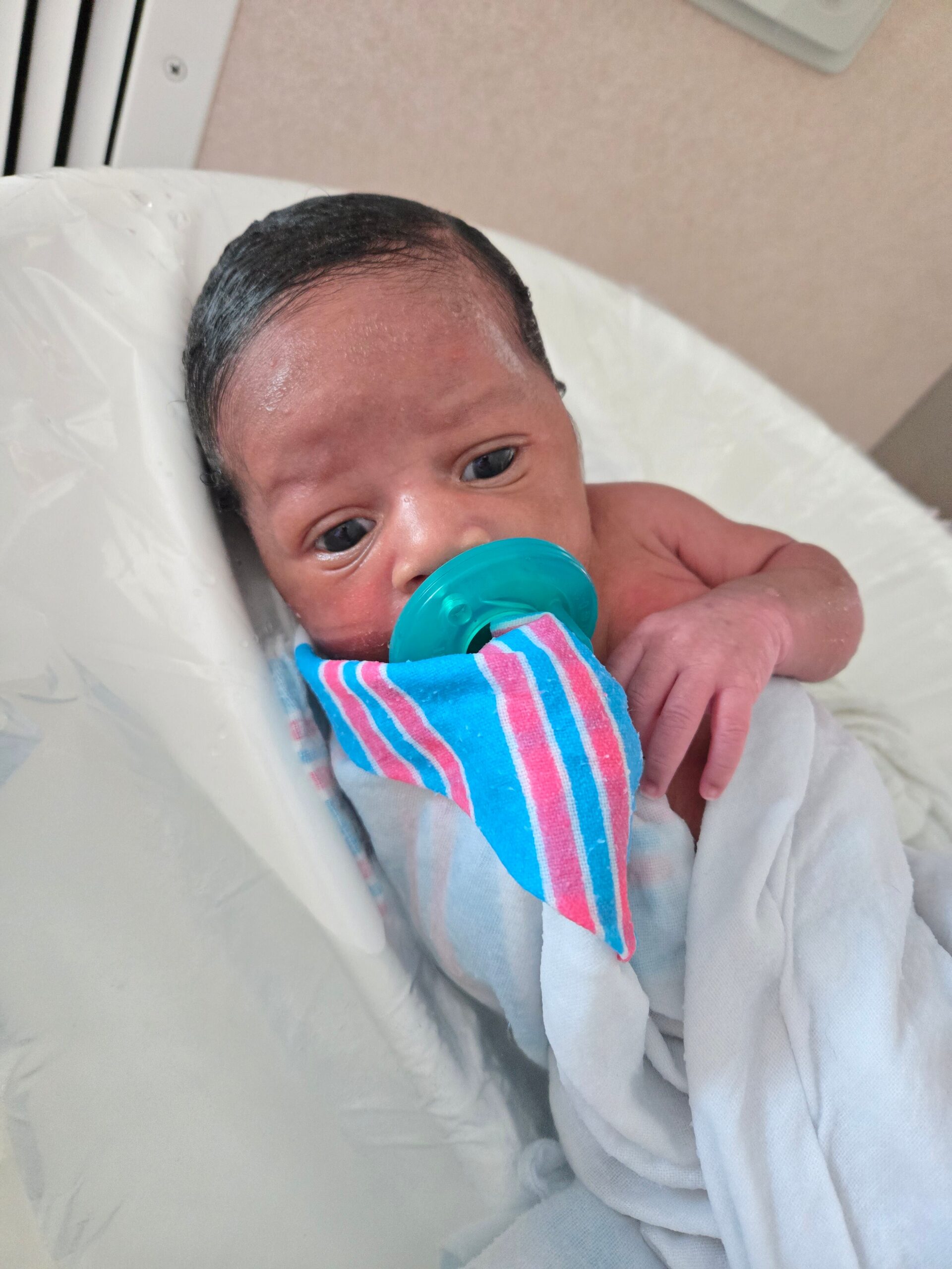 Newborn baby with dark hair, pacifier in mouth, wrapped in a blue-pink striped blanket on a hospital bed.
