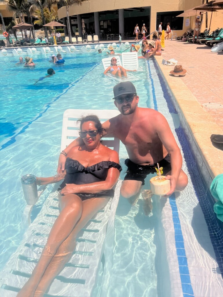 Couple lounging on pool chairs in a resort pool; woman in a black swimsuit with sunglasses, man shirtless with a cap holding a coconut drink.