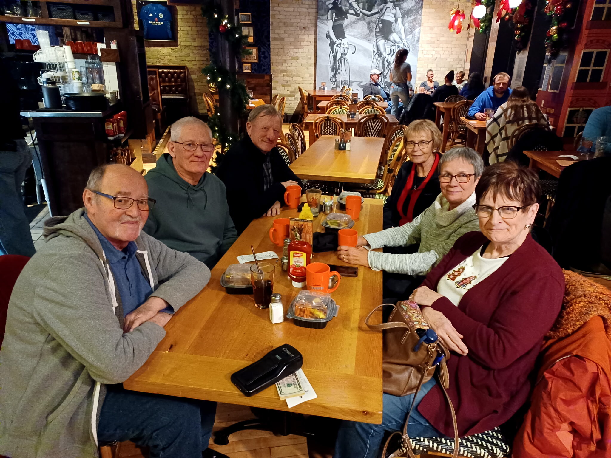 Group of older adults seated around a wooden table in a cozy cafe, with orange mugs and condiments, Christmas décor in the background.