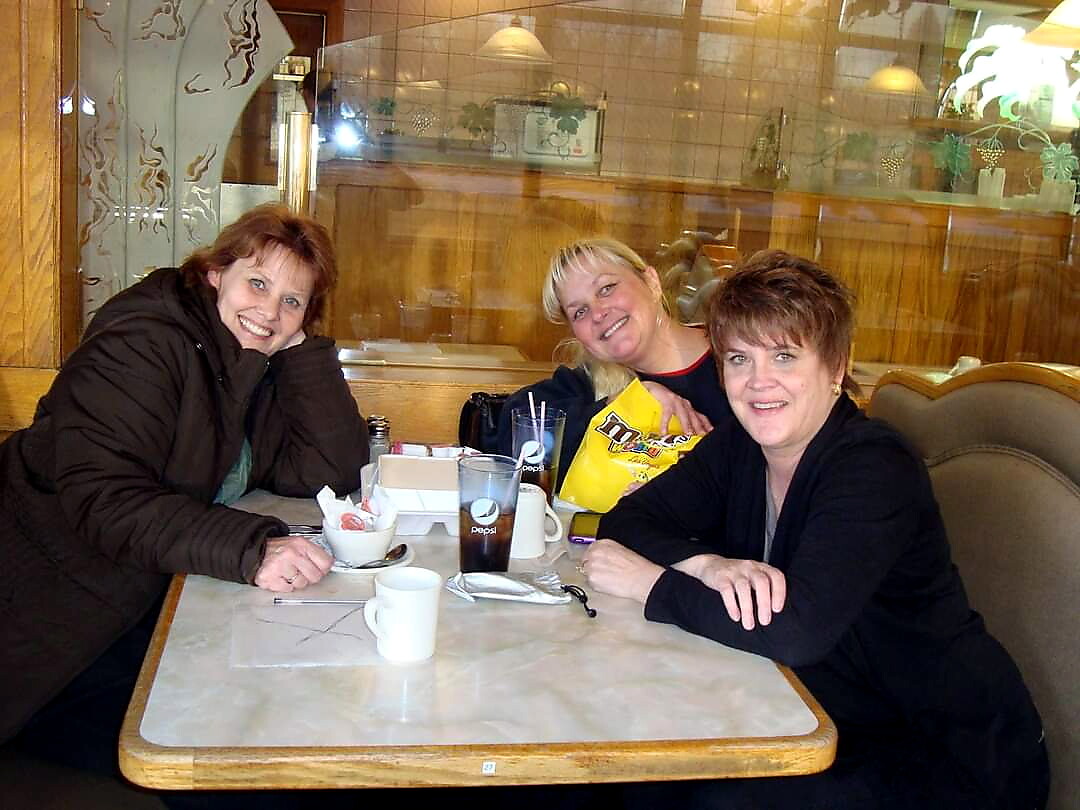 Three women sit at a cafe table, smiling at the camera; one holds a yellow bag of M&M's snacks in her lap.