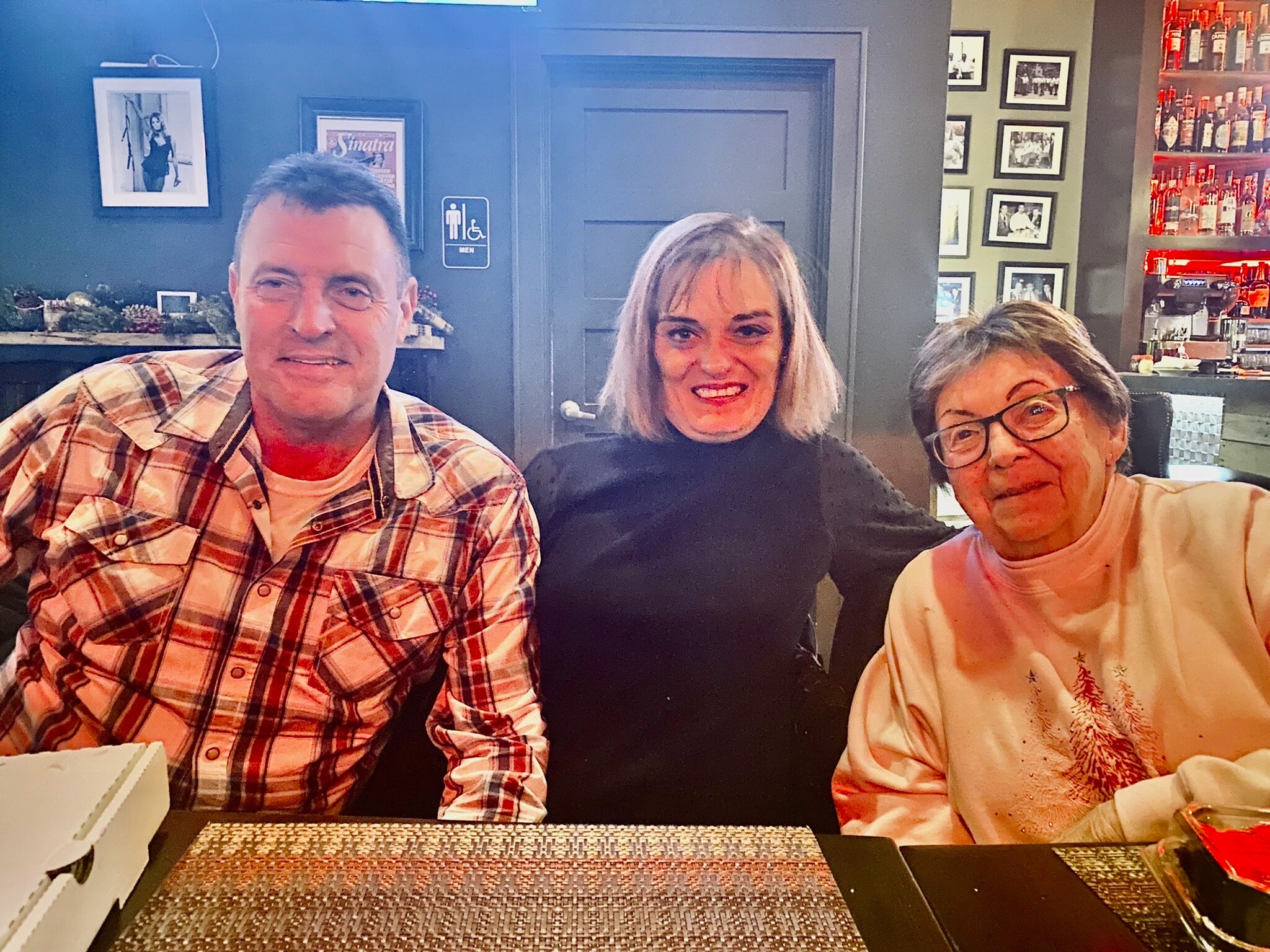 Three people smile at the camera while seated at a restaurant table: a man in a plaid shirt on the left, a woman with light hair in the middle, and a woman with glasses on the right, with a bar and photos in the background.