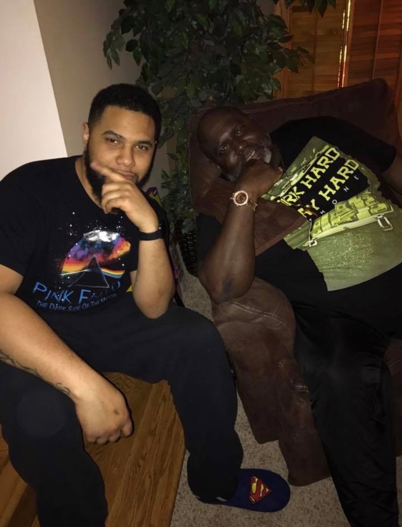 Two men sit together on a sofa indoors; the younger man on the left rests his chin on his hand, wearing a black Pink Floyd shirt, while the older man on the right leans back in a brown jacket with a gold watch.