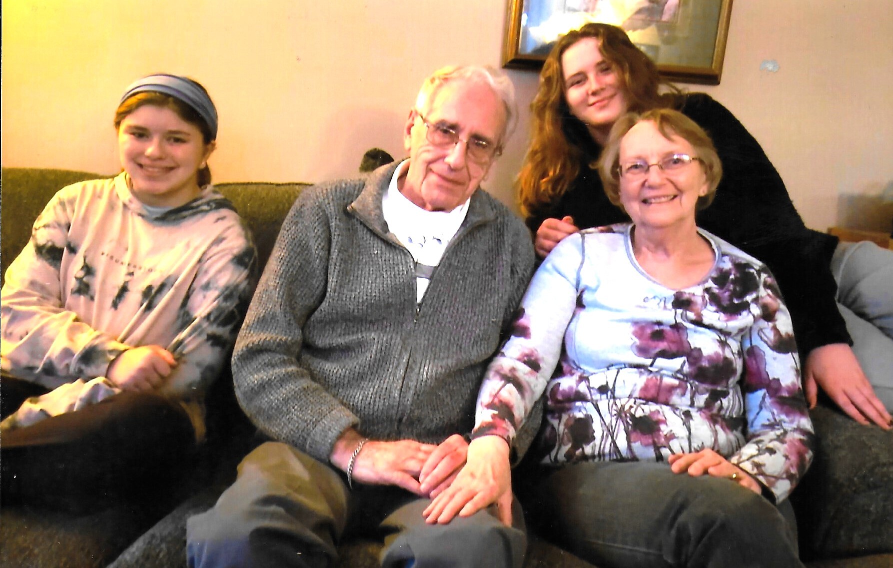 Four people sitting on a sofa: a young girl with a blue headband, an elderly man in a gray sweater, and two smiling women—one in a floral top and another behind them.