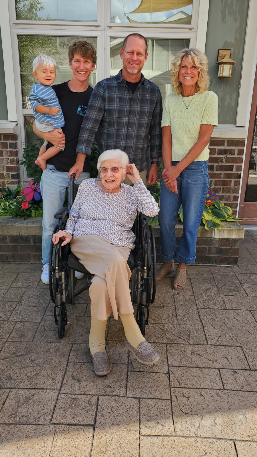 Smiling family posing outdoors in front of a brick house with flowers; elderly woman in a wheelchair, flanked by three adults and a toddler.