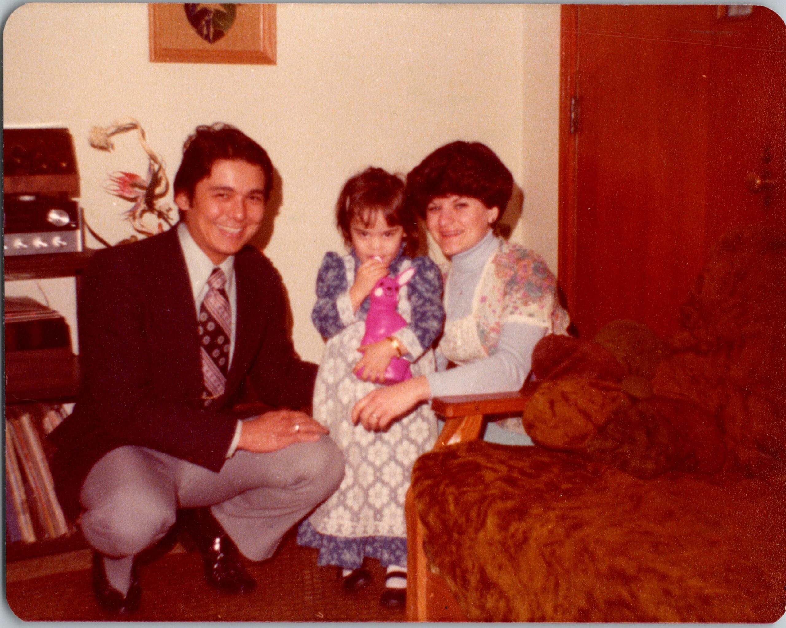 Family trio smiling for a 1970s-style indoor photo: man in suit, woman, and child holding a pink stuffed animal.
