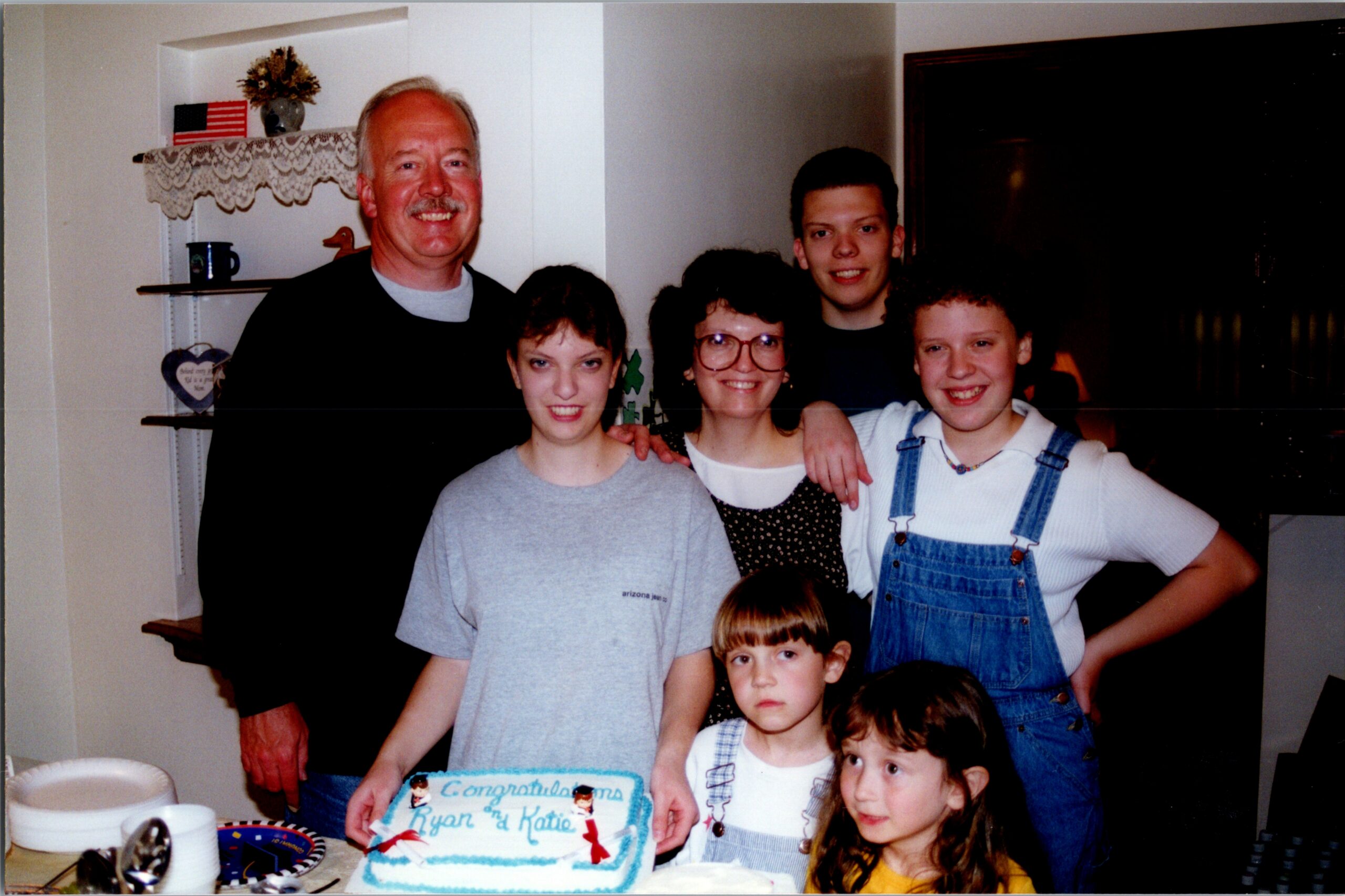 Group of adults and children posing indoors around a birthday cake on a table. The scene is a casual family celebration with smiles and decorations nearby.