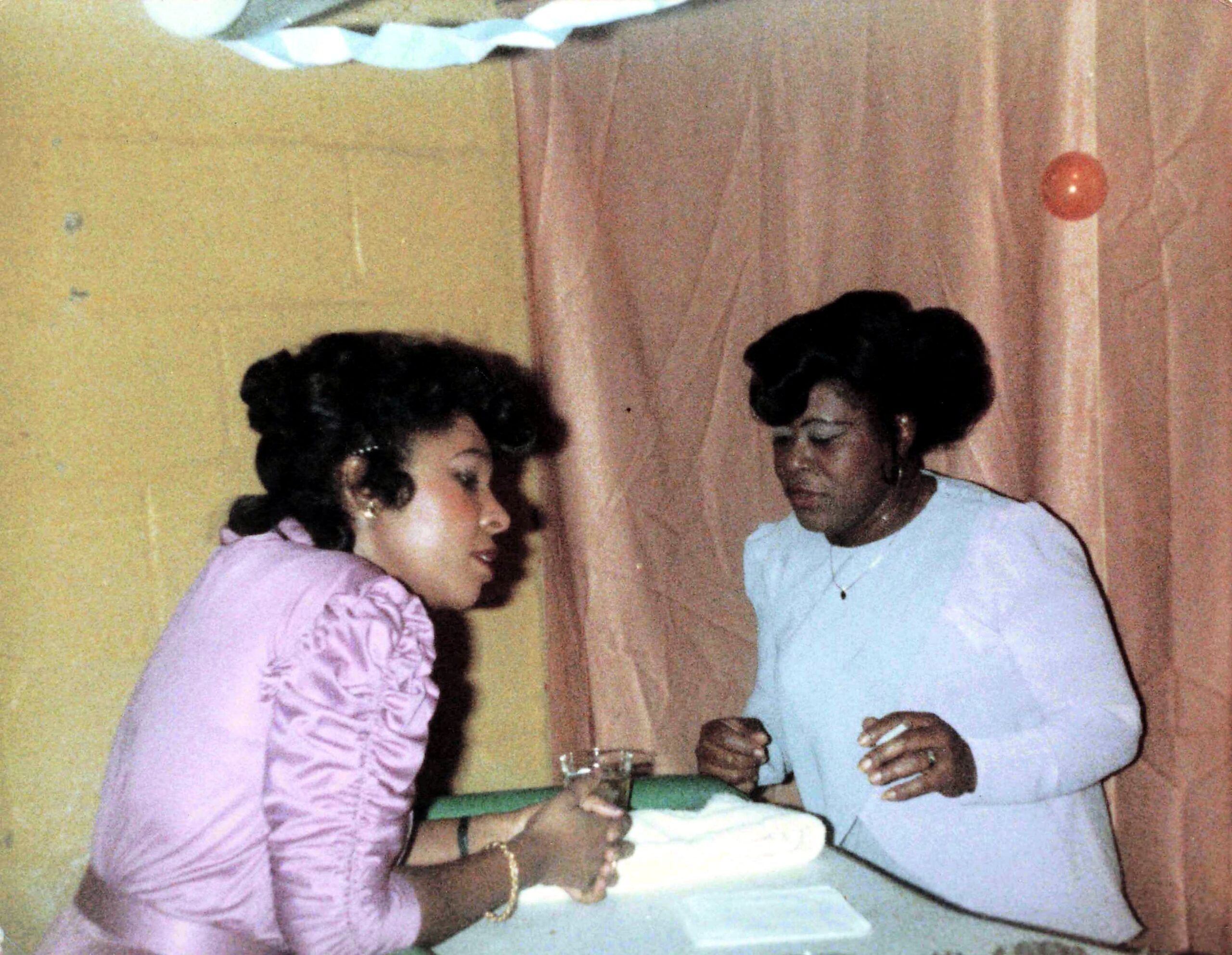 Two women in pastel dresses stand at a table cutting a cake together at a celebration, with a pink curtain and a red balloon in the background.