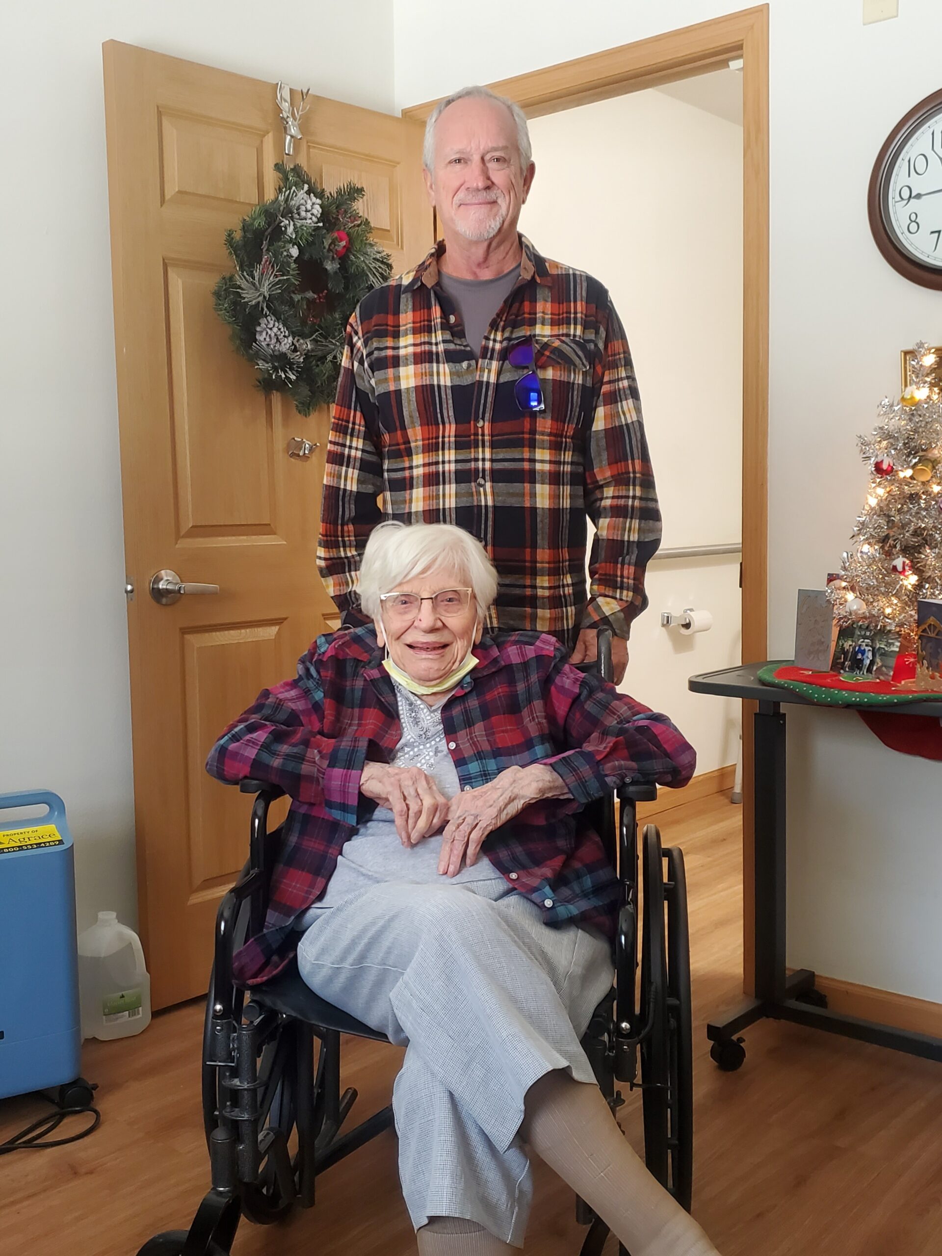 Smiling elderly woman in a wheelchair in the foreground; man in a plaid shirt stands behind her, with a holiday wreath on the door and a Christmas tree nearby.