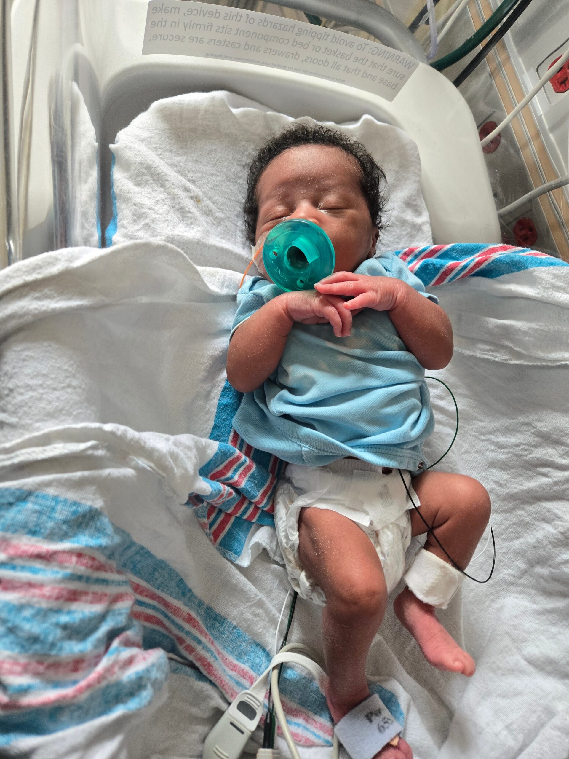Newborn baby lying in a hospital incubator with a teal pacifier, connected to monitoring wires and tubes, wrapped in a blue outfit and blankets.