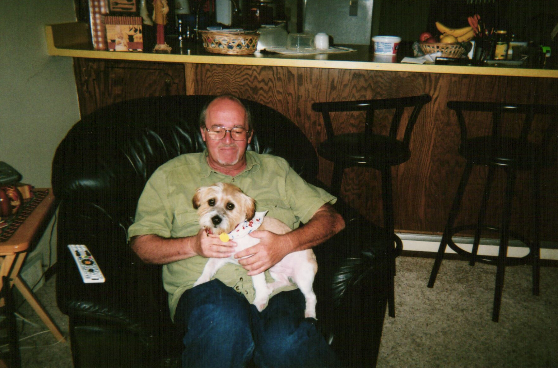 Older man with glasses sitting in a black leather chair, holding a small white dog wearing a bandana.