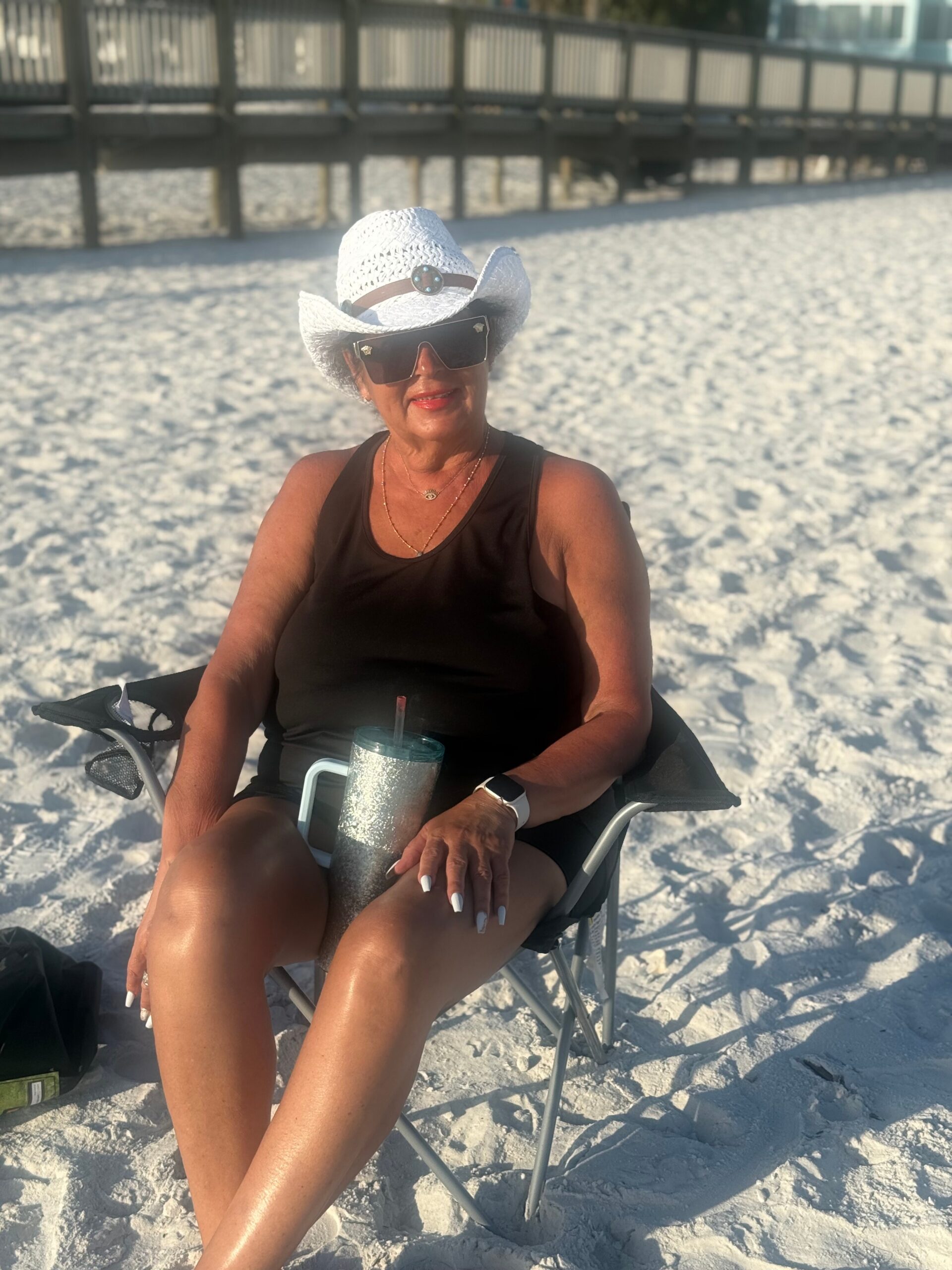 Woman in a white cowboy-style hat and oversized sunglasses sits on a beach chair, holding a glittery tumbler on the sand.