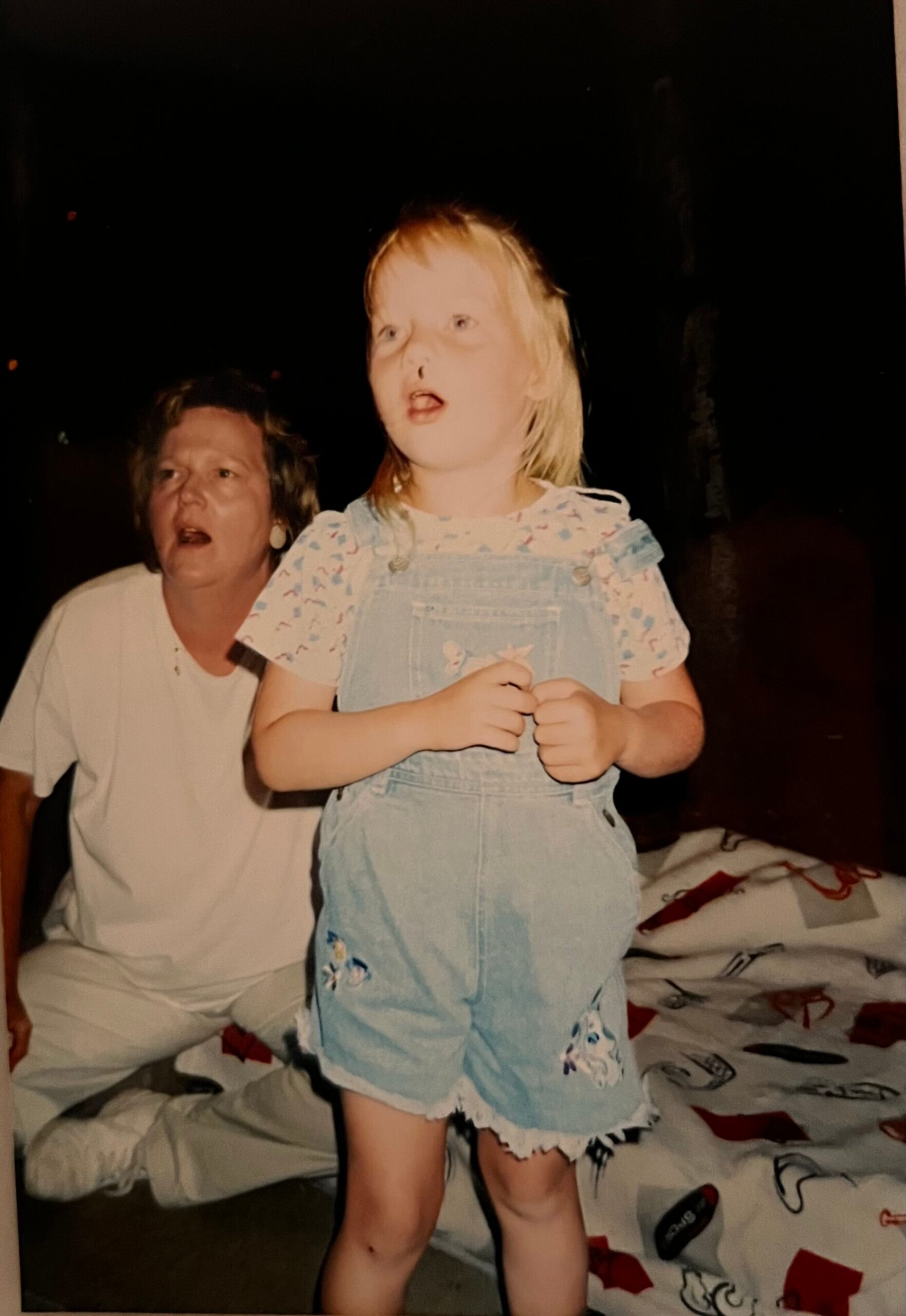 Young girl in light-blue denim overalls and a patterned shirt stands in a dimly lit room with an adult behind her.