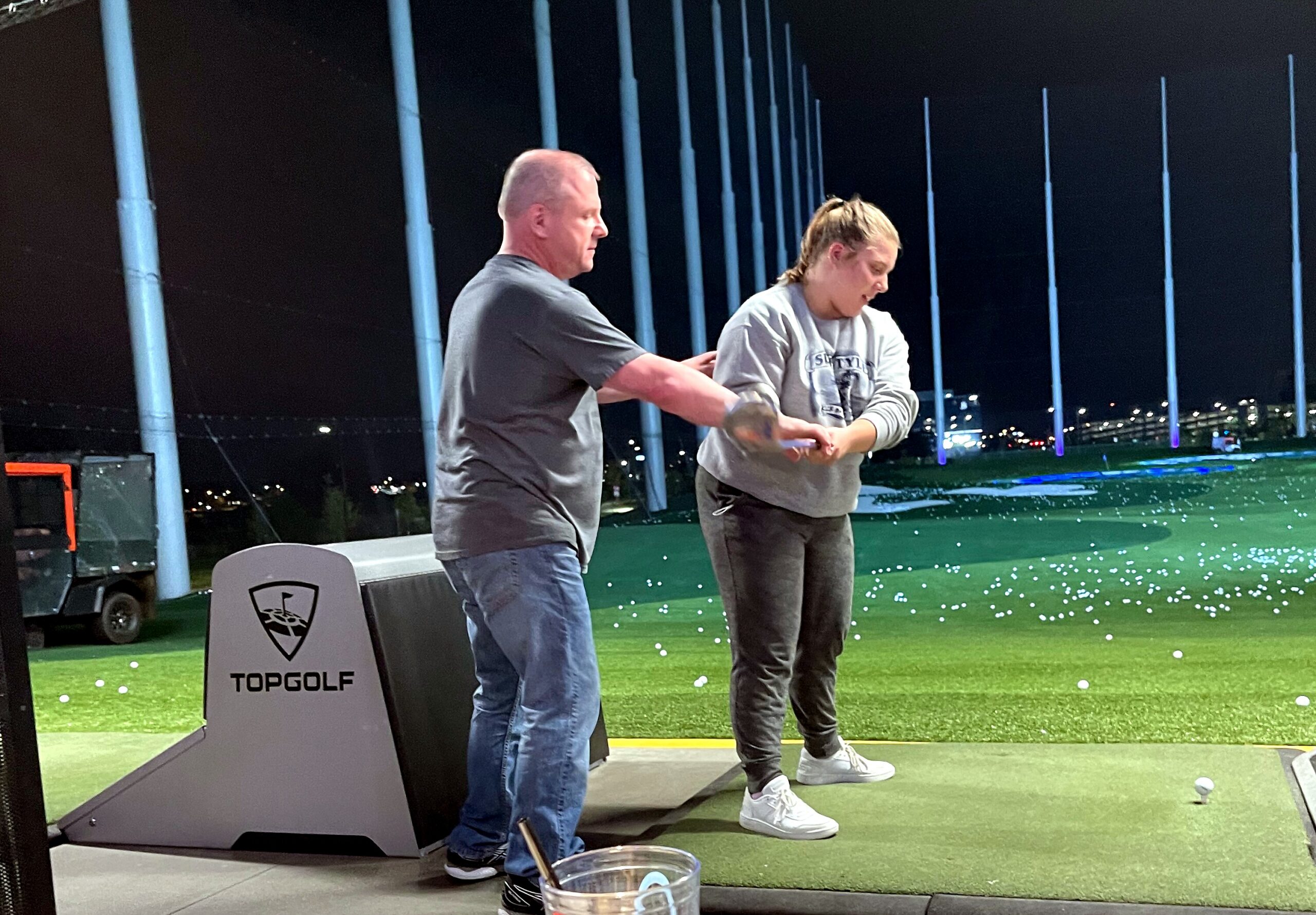 Two adults stand on a practice mat at Topgolf at night, with golf balls scattered around and a man guiding a woman.