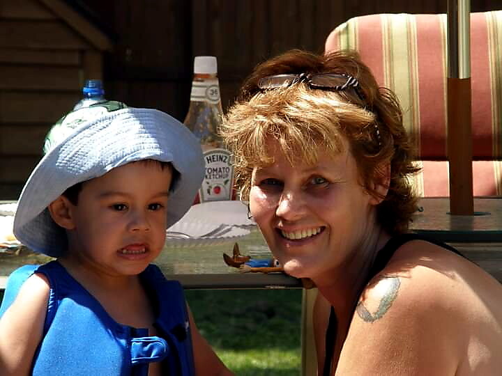Smiling woman with short hair and sunglasses beside a young boy in a blue outfit and light-blue hat outdoors, with a patio table in the background.
