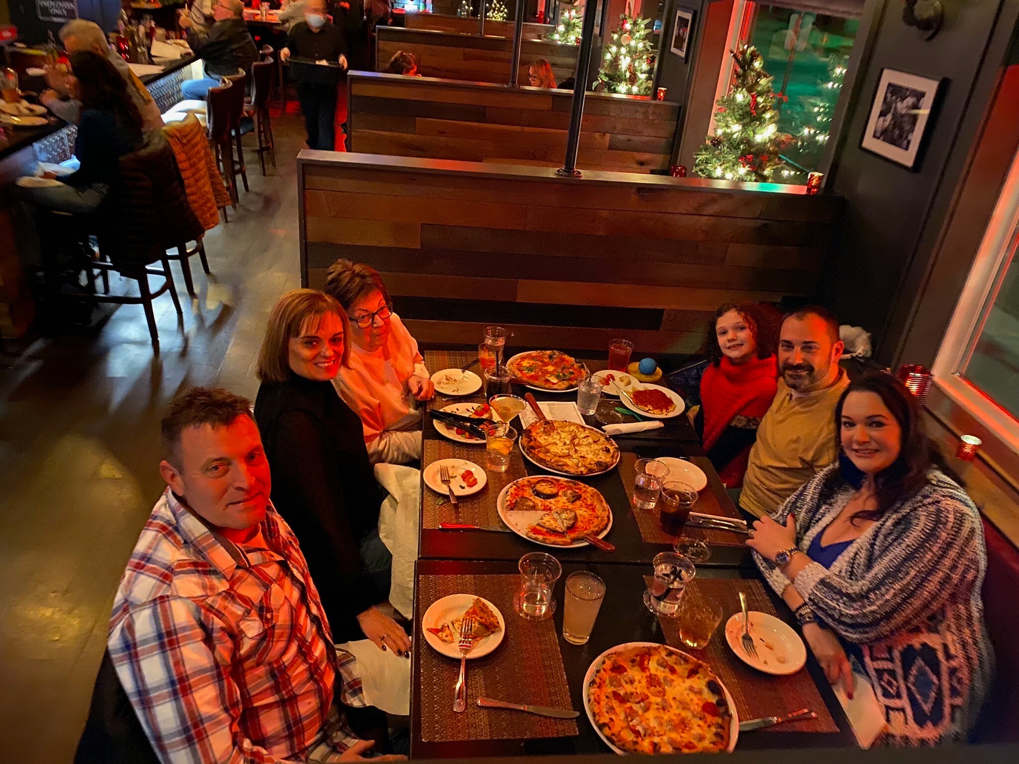 Group of six to eight people around a dining table with pizzas, drinks, and plates in a cozy, festively lit restaurant.
