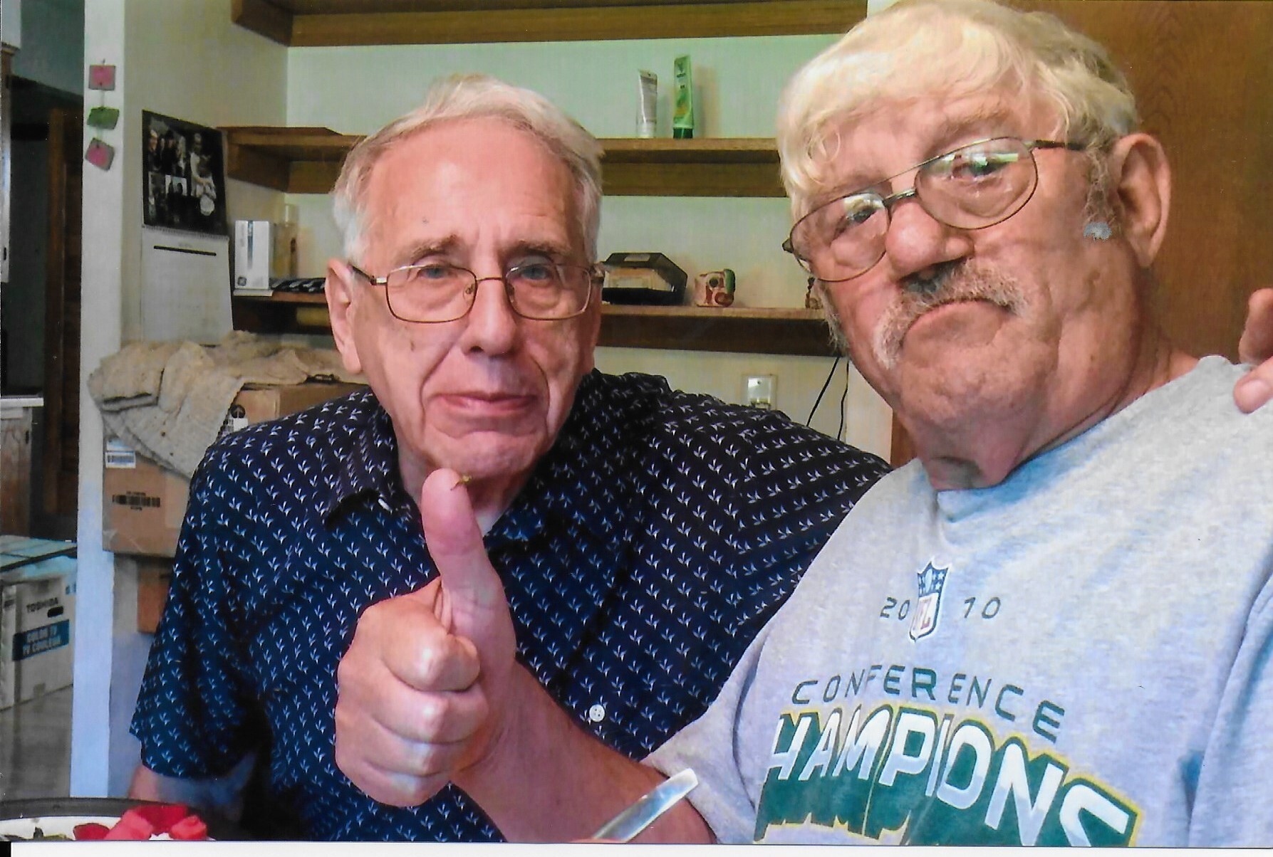 Two smiling older men take a selfie indoors, one in a dark patterned shirt giving a thumbs up toward the camera.