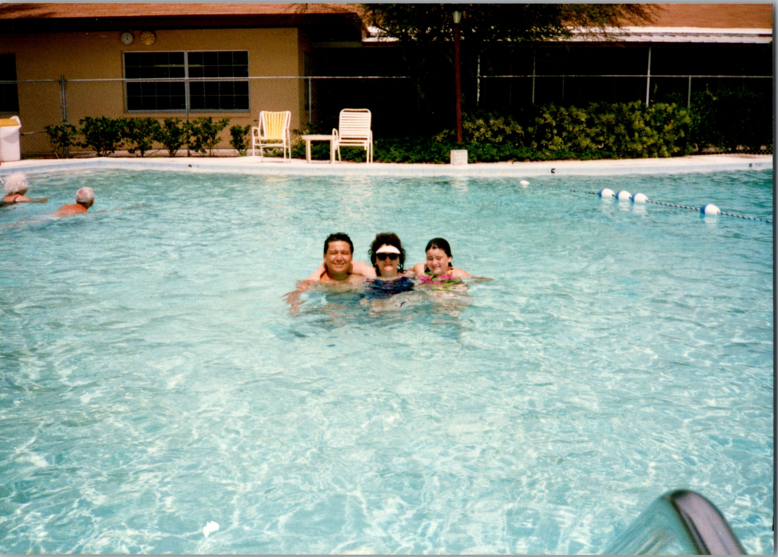 Three people swim in a bright outdoor pool, facing the camera with the building and lounge chairs in the background.