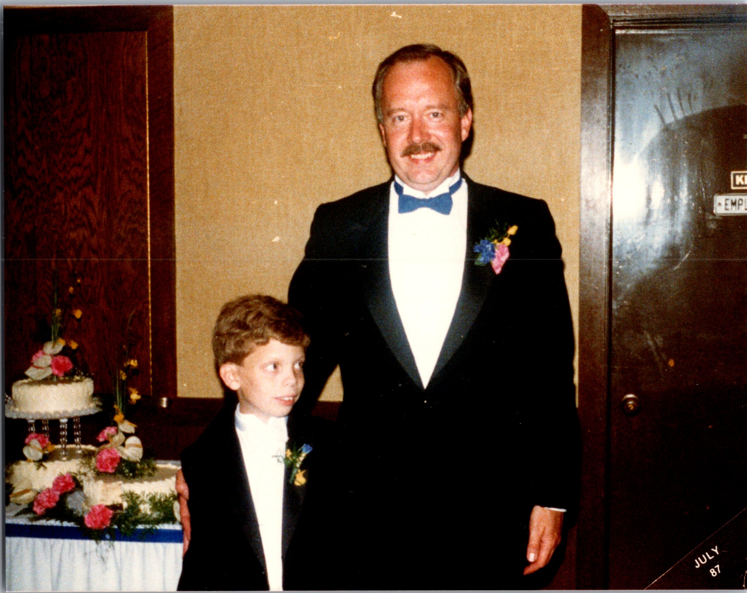Man in formal tuxedo with blue bow tie posing with a boy in a tuxedo at a wedding, cake and flowers in the background