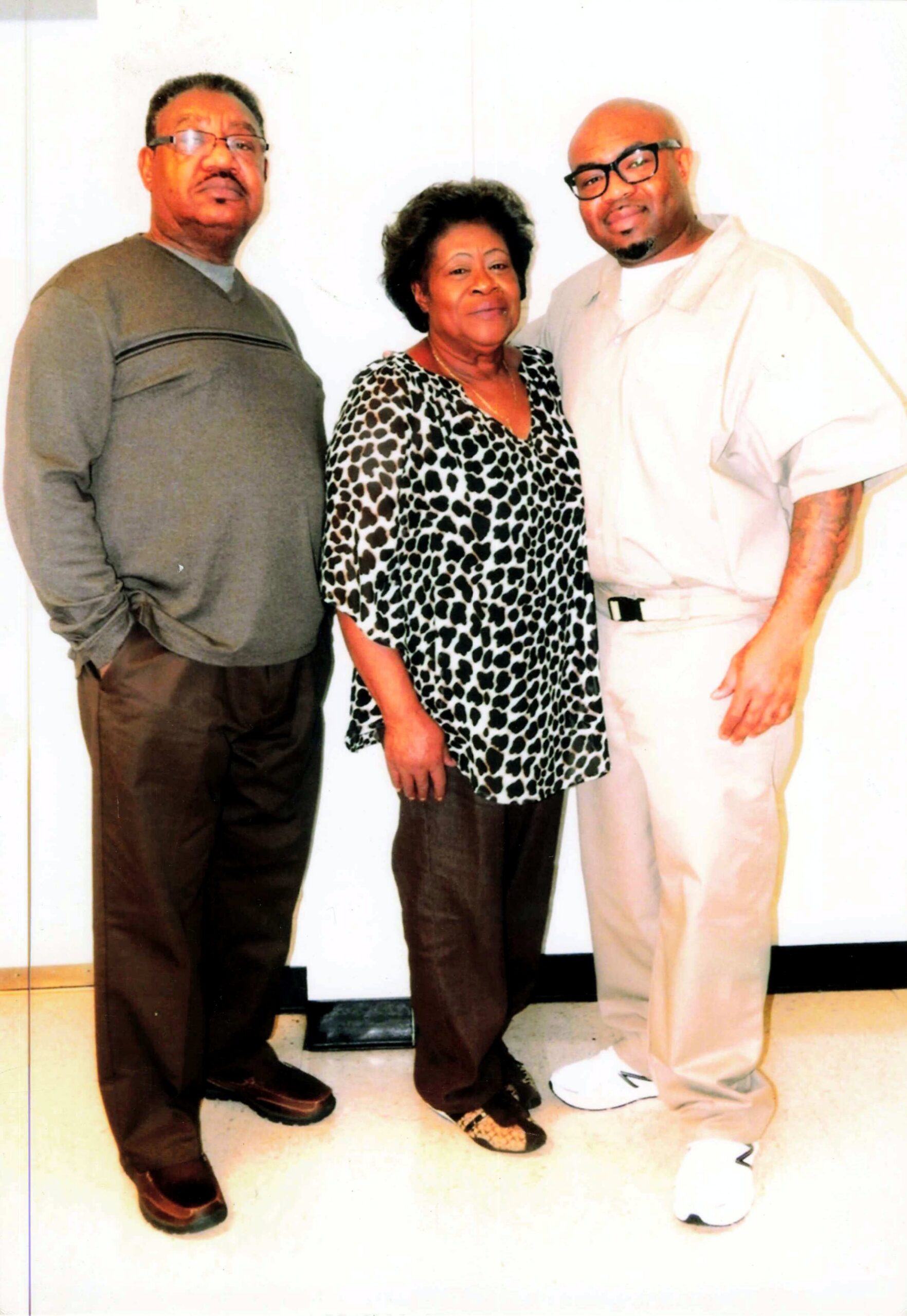 Three adults posing together indoors: a man in a gray sweater on the left, a woman in a leopard-print blouse in the center, and a man in a light beige suit with glasses on the right, all smiling slightly.