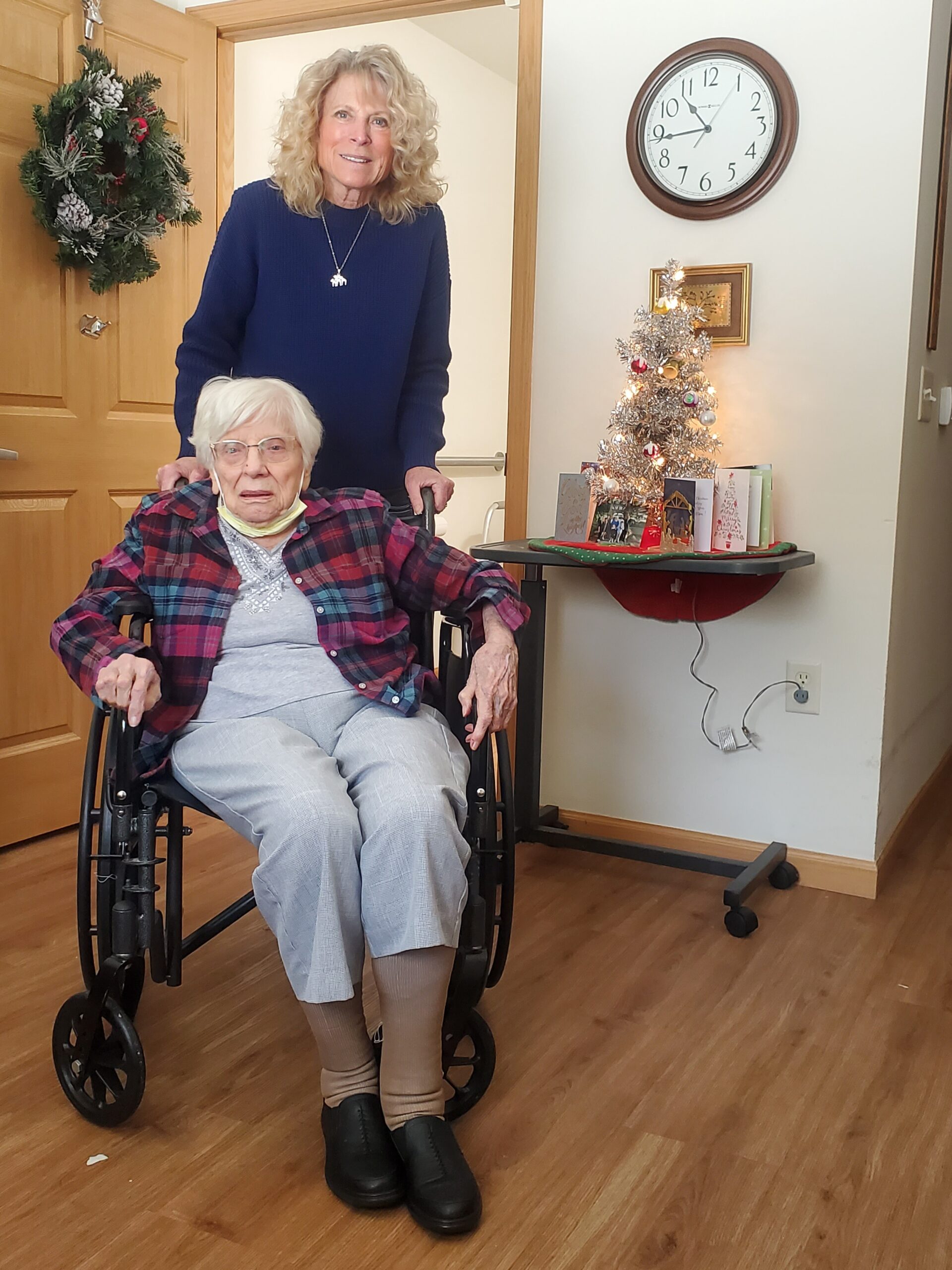 Older woman in a wheelchair with a caregiver standing behind her in a Christmas-decorated living room.