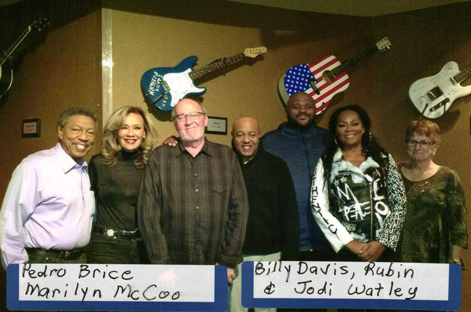 Six people standing together in a venue with guitars on the wall behind them, smiling for the photo; signs display names including Pedro Brice, Marilyn McCoo, Billy Davis, Rubin, and Jodi Watley.