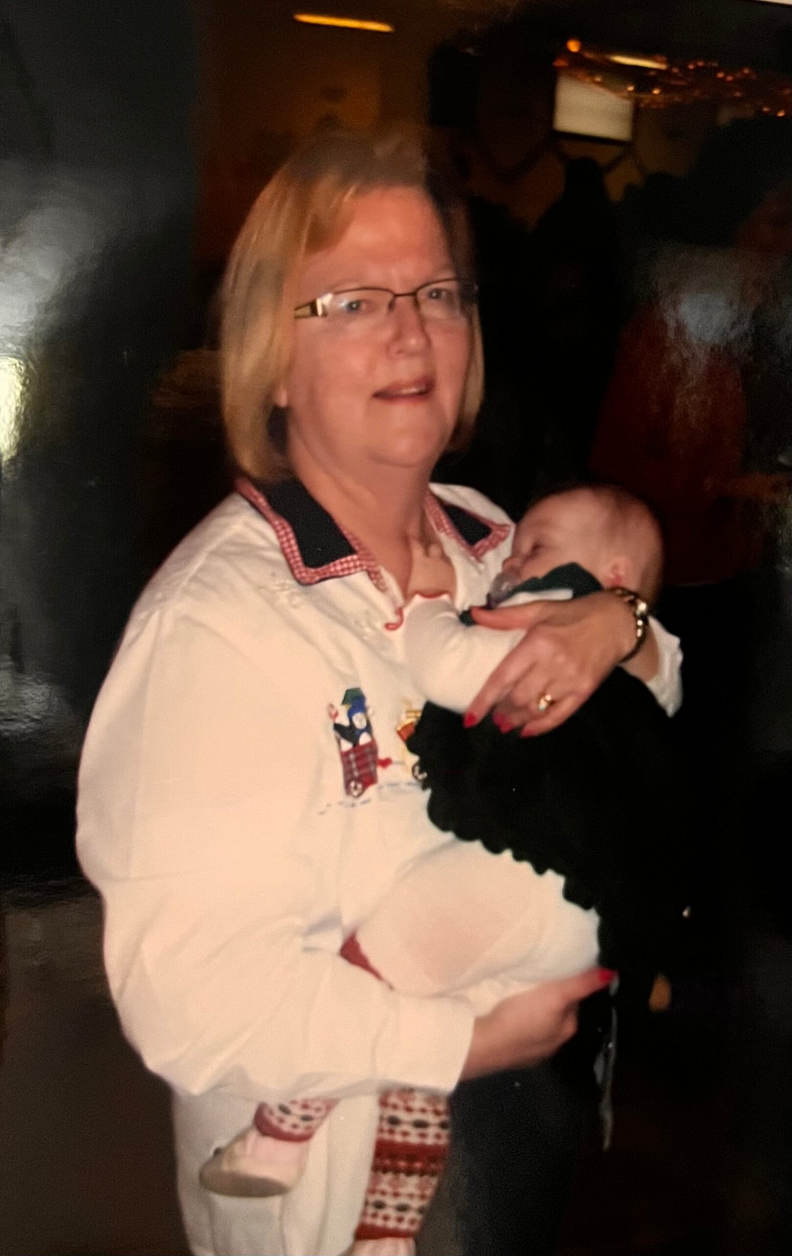 Woman with short blond hair and glasses cradling a sleeping baby in her arms at a dim event venue.