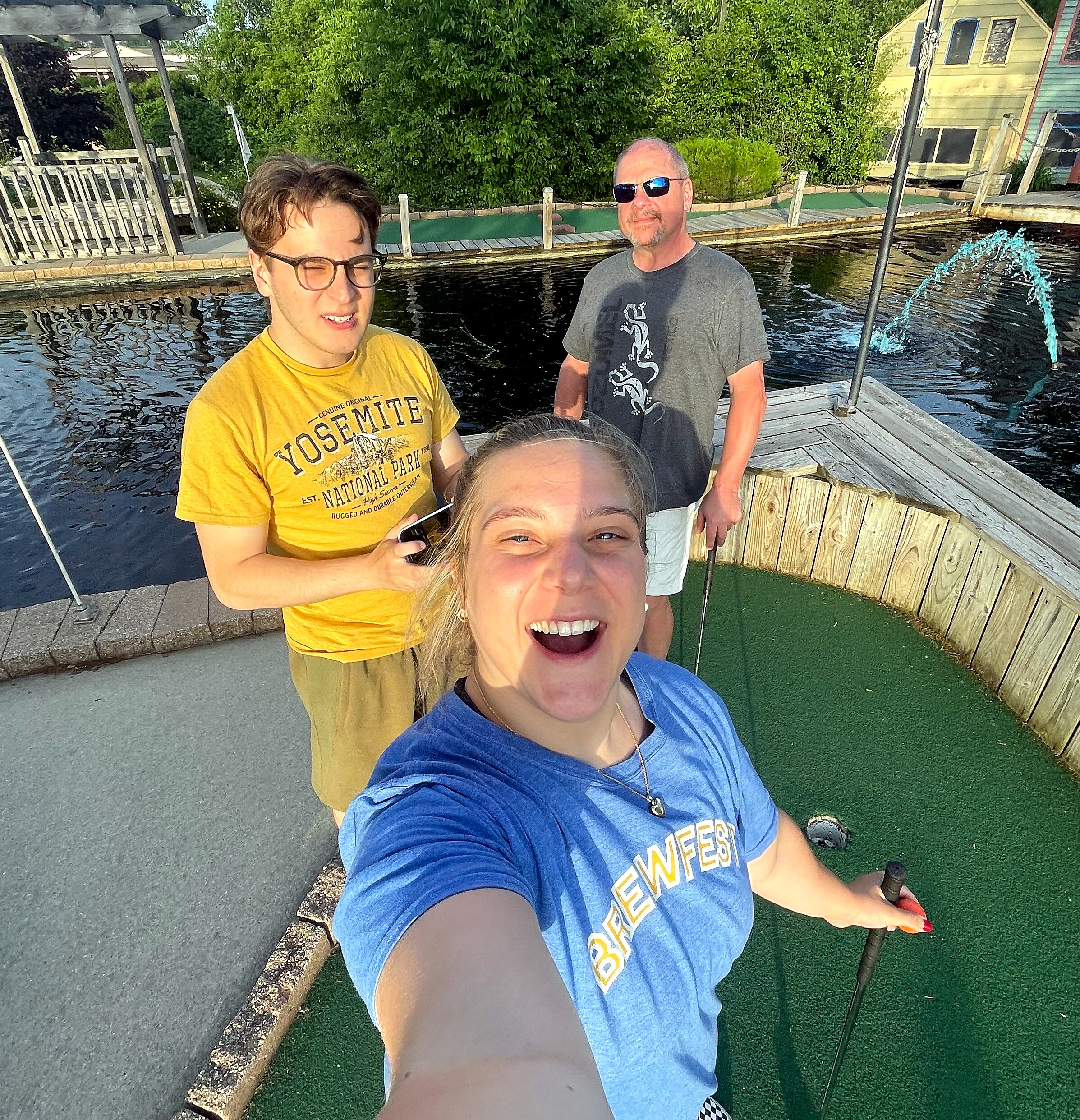 Group selfie on a wooden deck beside a pool of water with a fountain, smiling people in casual shirts (yellow and blue).
