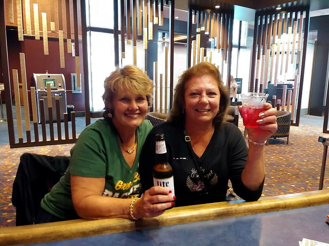 Two smiling women sit at a bar, each holding a drink—a beer bottle and a red cocktail in a plastic cup.