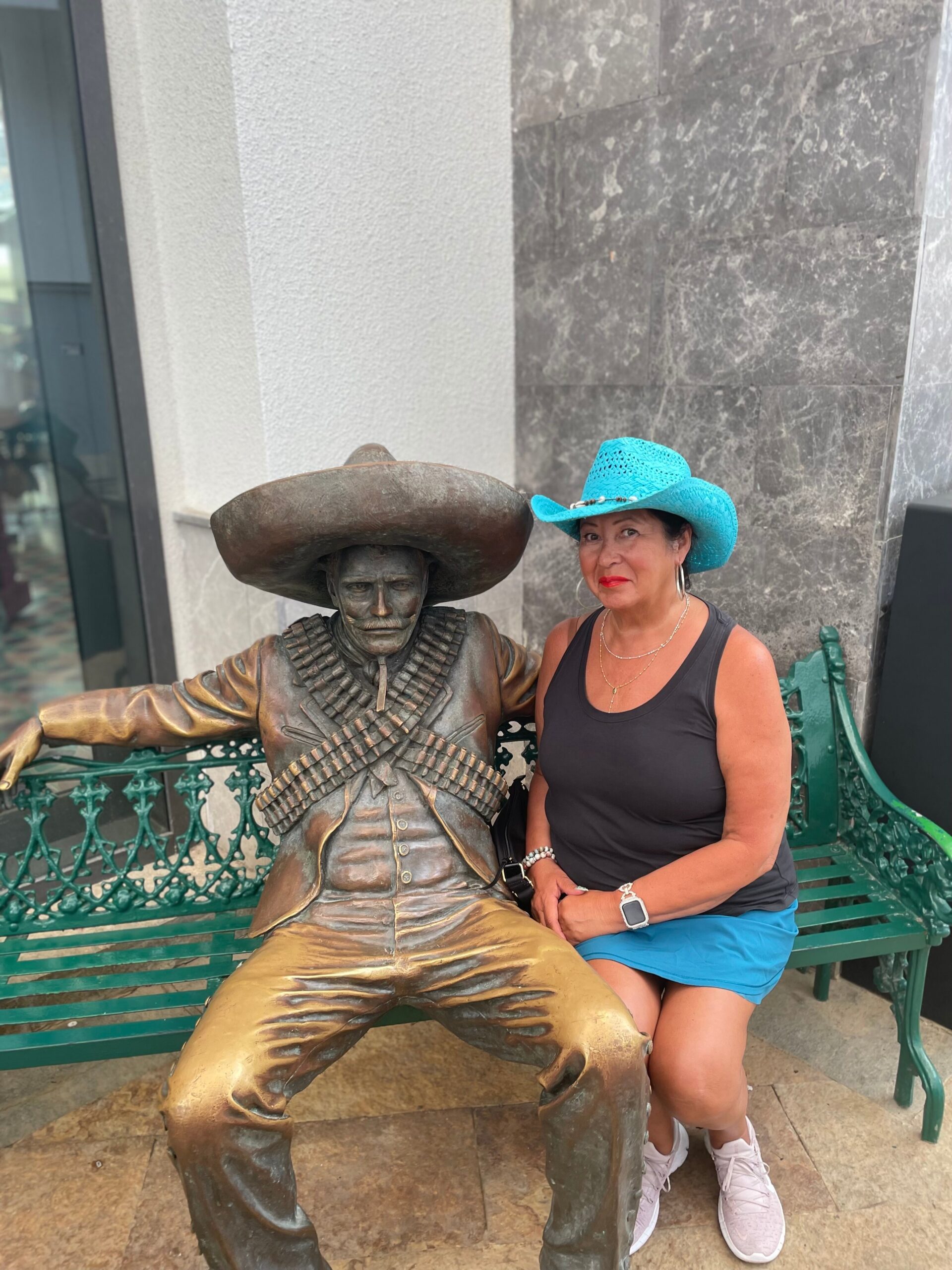 Woman wearing a turquoise cowboy hat sits on a green bench beside a bronze statue of a man in a large sombrero.