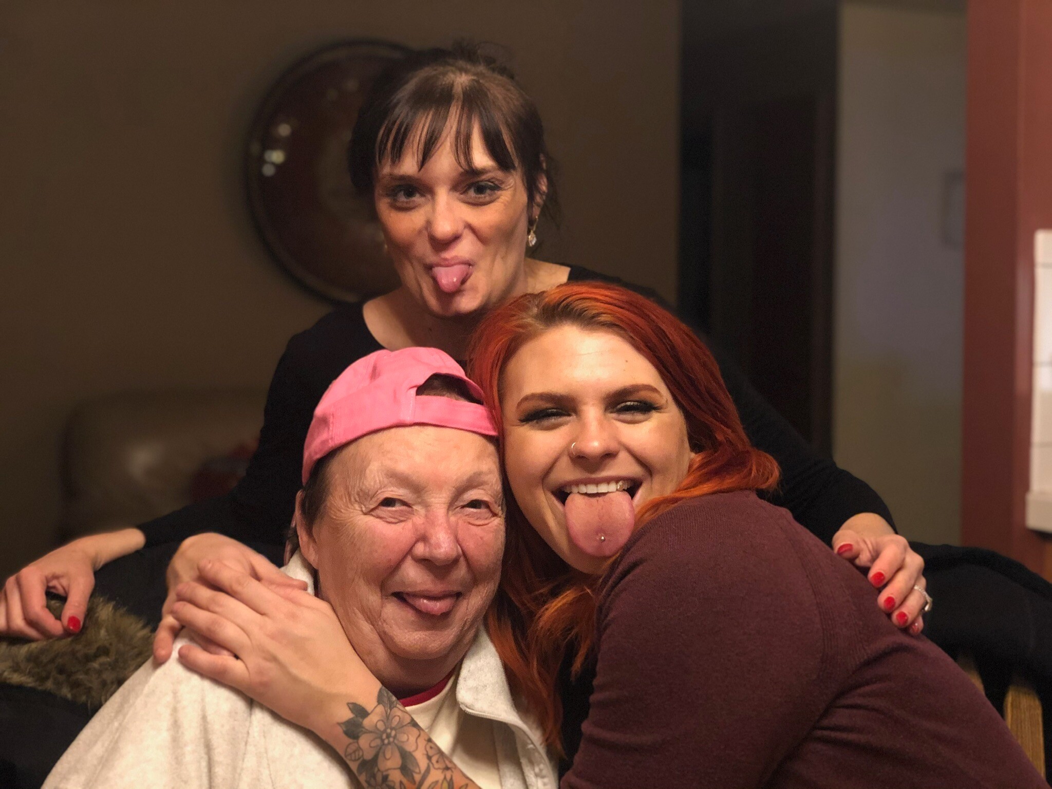 Three friends posing closely indoors, smiling and sticking out their tongues in a playful selfie.