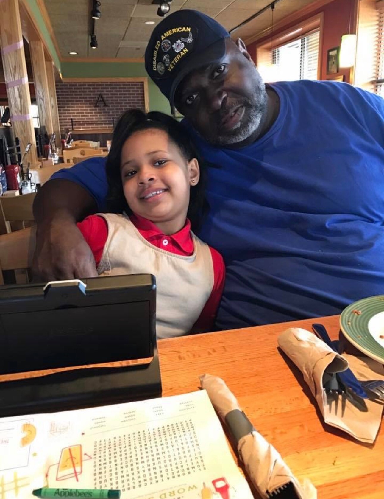 Man in a blue shirt wearing a veteran cap sits with a smiling girl at a restaurant table, his arm around her.